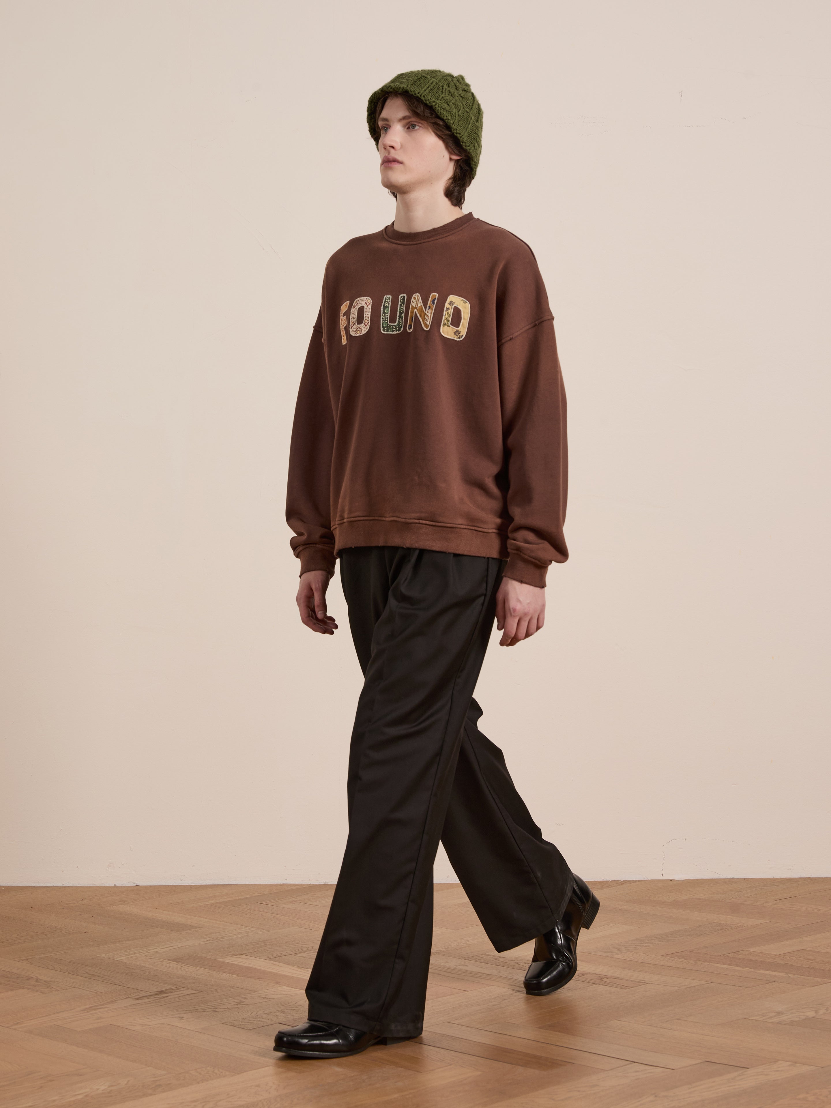 A person wearing a FOUND brand Patterned Logo Crewneck sweater, black pants, black shoes, and a green beanie strolls across the wooden floor.