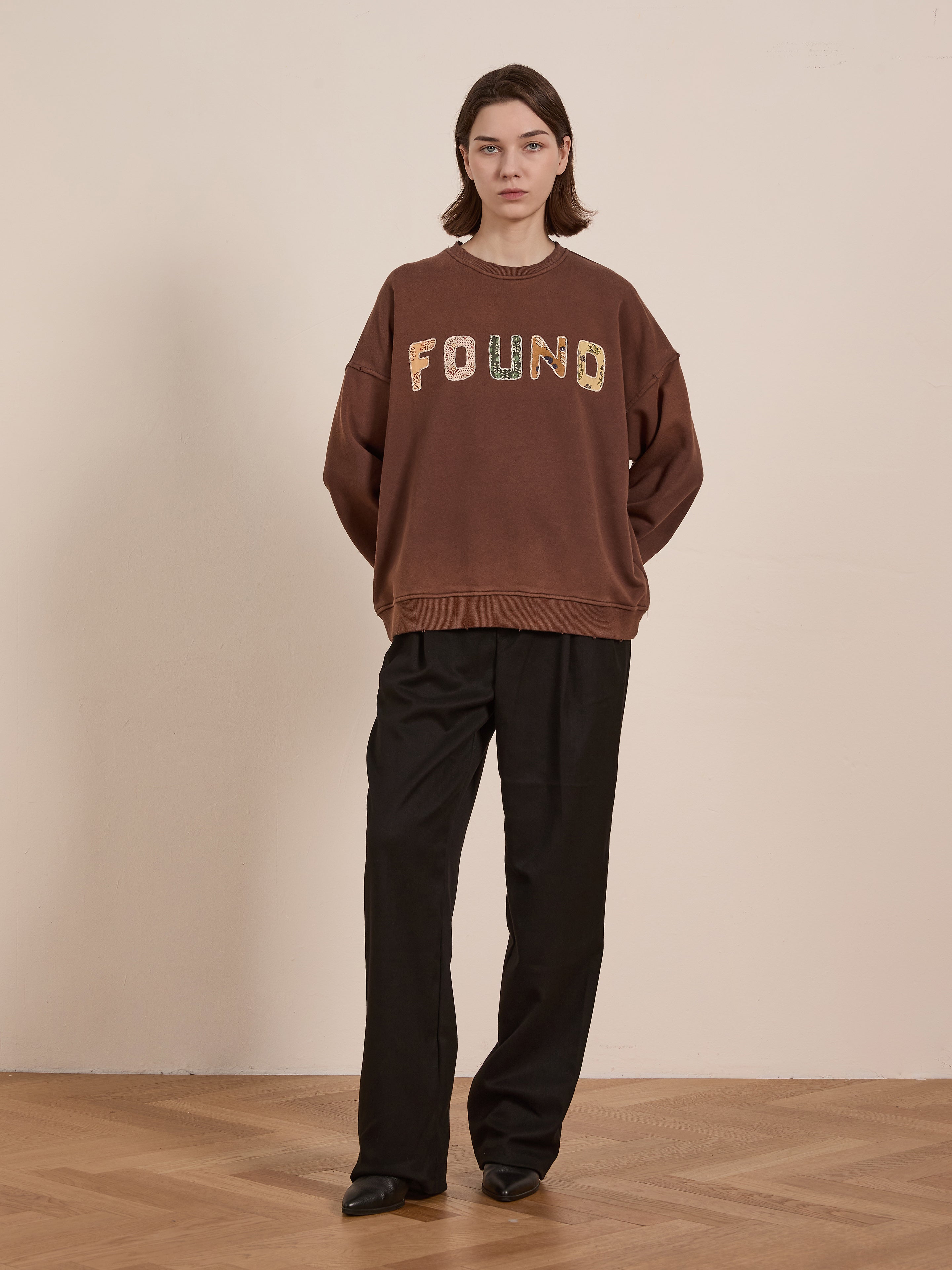 A person stands against a plain background wearing the FOUND Patterned Logo Crewneck in brown french terry cotton, paired with black pants and black shoes.