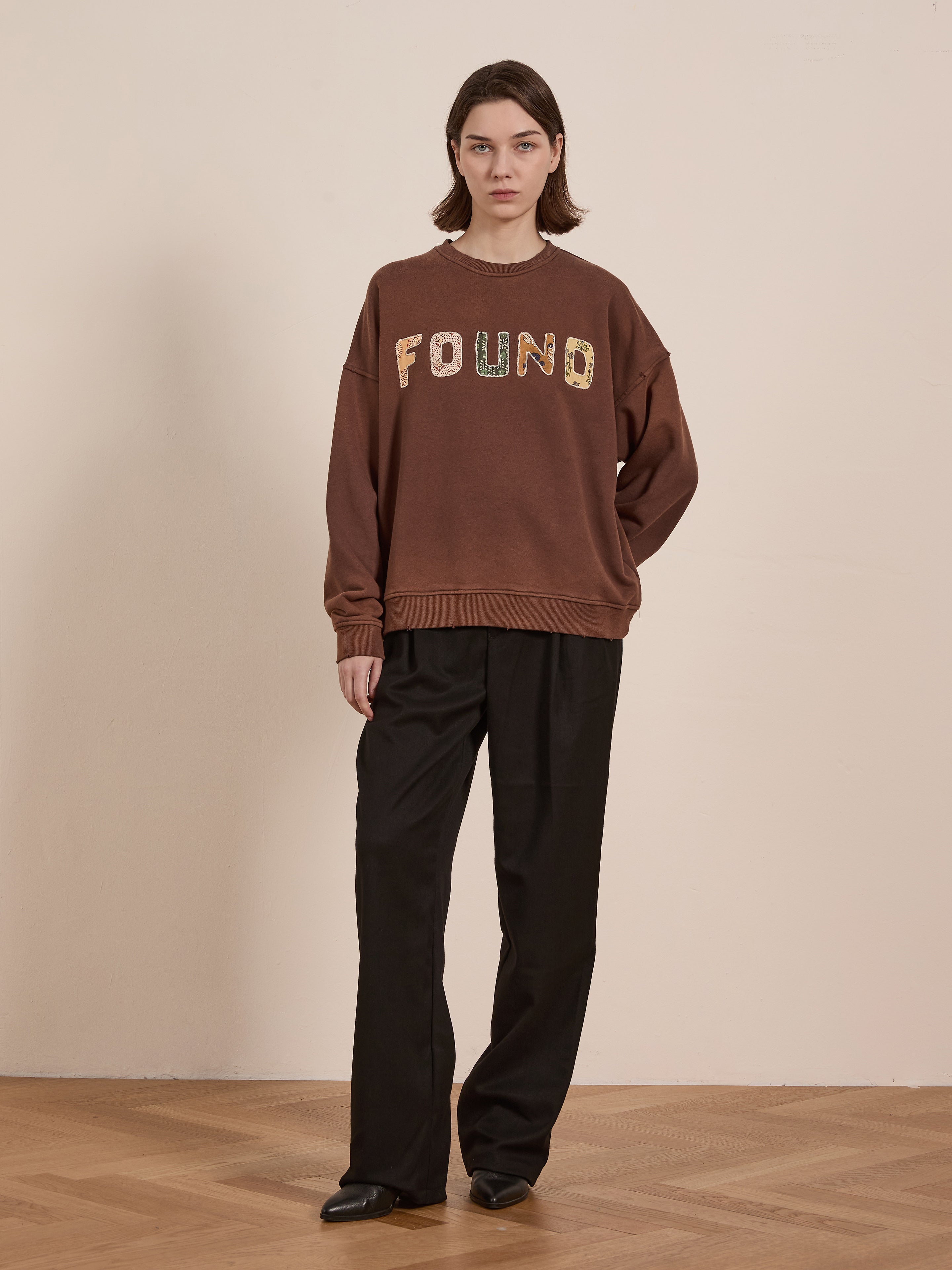 Indoors on a wooden floor, a person wears FOUND's Patterned Logo Crewneck in brown French terry cotton with appliqué "FOUND" lettering and black pants, set against a beige wall backdrop.