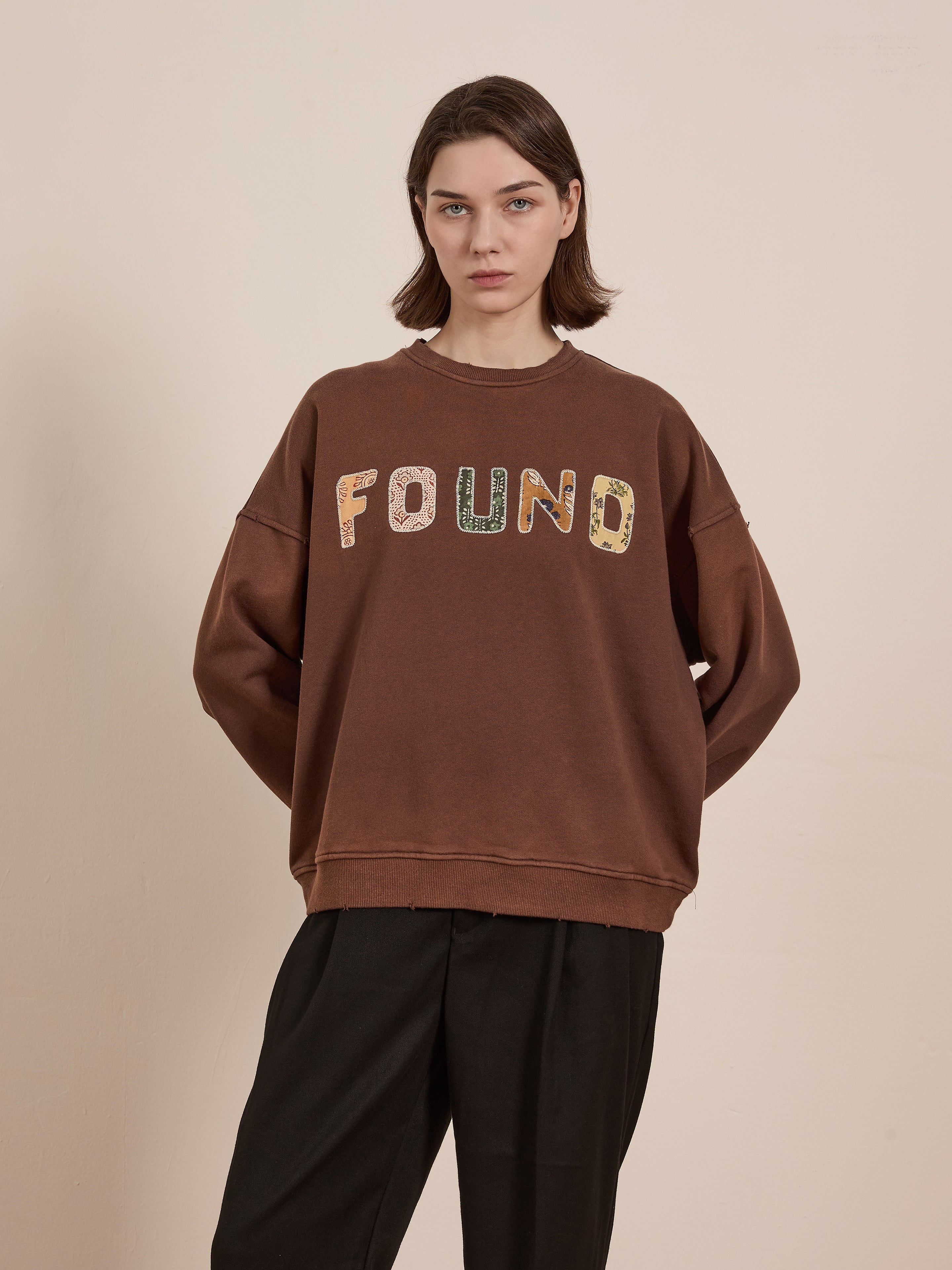 A person stands against a beige background wearing the FOUND Patterned Logo Crewneck, featuring appliqué "FOUND" lettering.