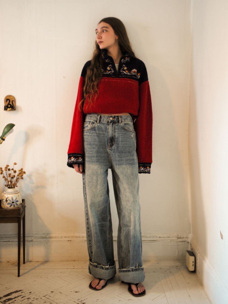 A person stands against a plain wall wearing a red and black sweater, Lacy Baggy Jeans by Found, and sandals. A small table with a potted plant is to their left.