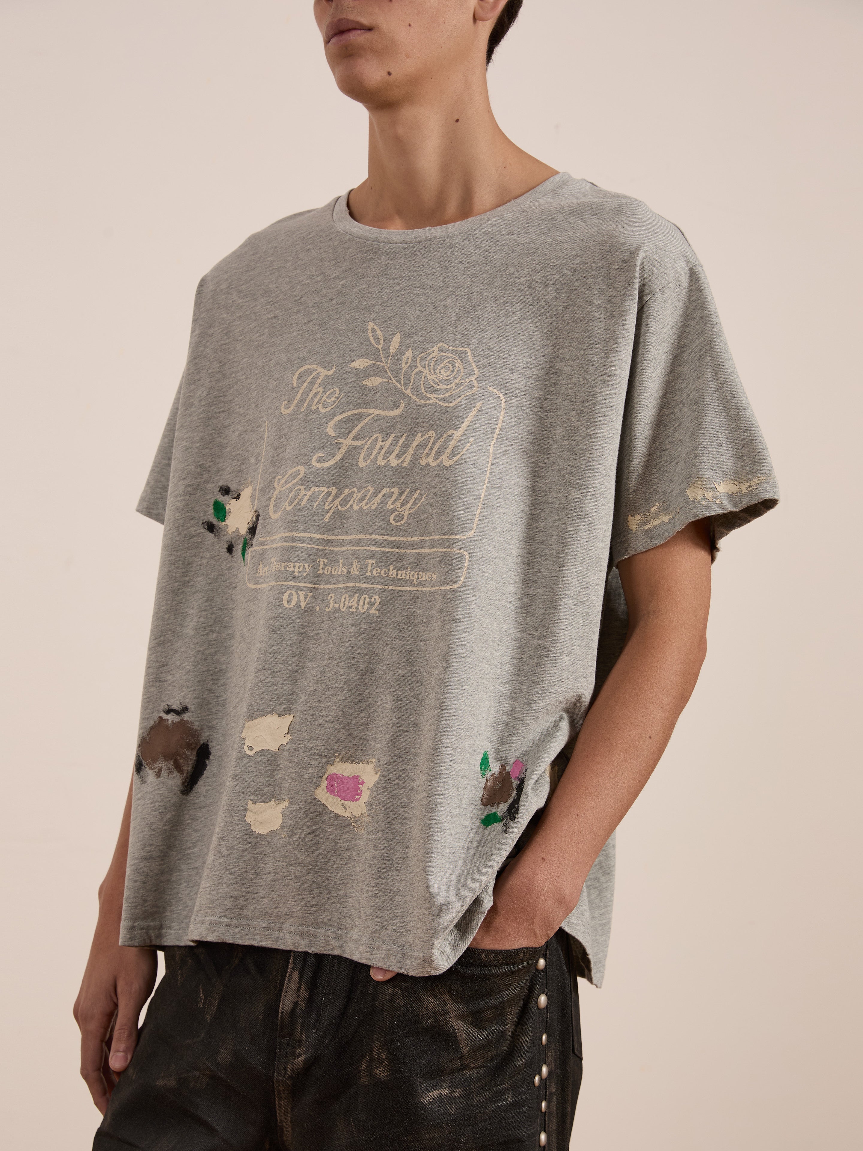 A person stands against a plain backdrop wearing a gray oversized "Painters Company Tee" by FOUND, featuring vintage-style paint splatters. Hands in pockets, the unisex look effortlessly embodies workwear-inspired style.