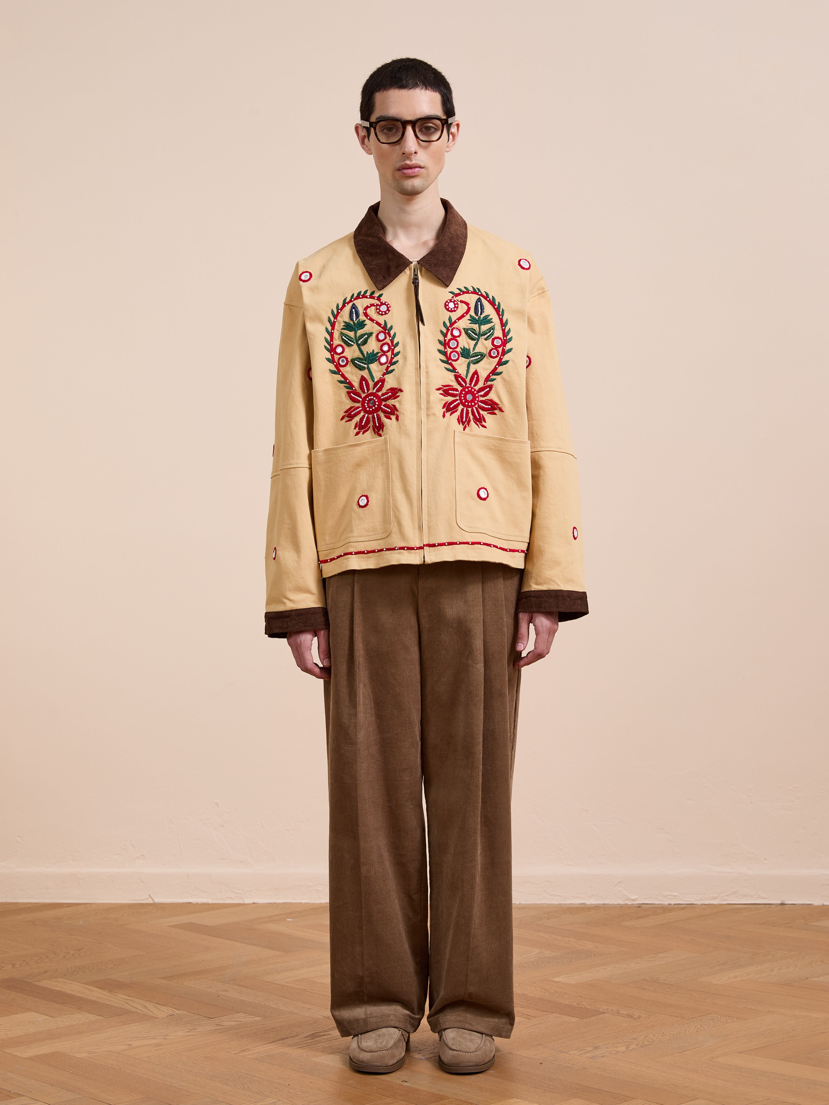 A person stands indoors against a plain wall, wearing glasses, the Found Paisley Motif Hand Embroidered Work Jacket, brown wide-leg pants, and tan shoes.