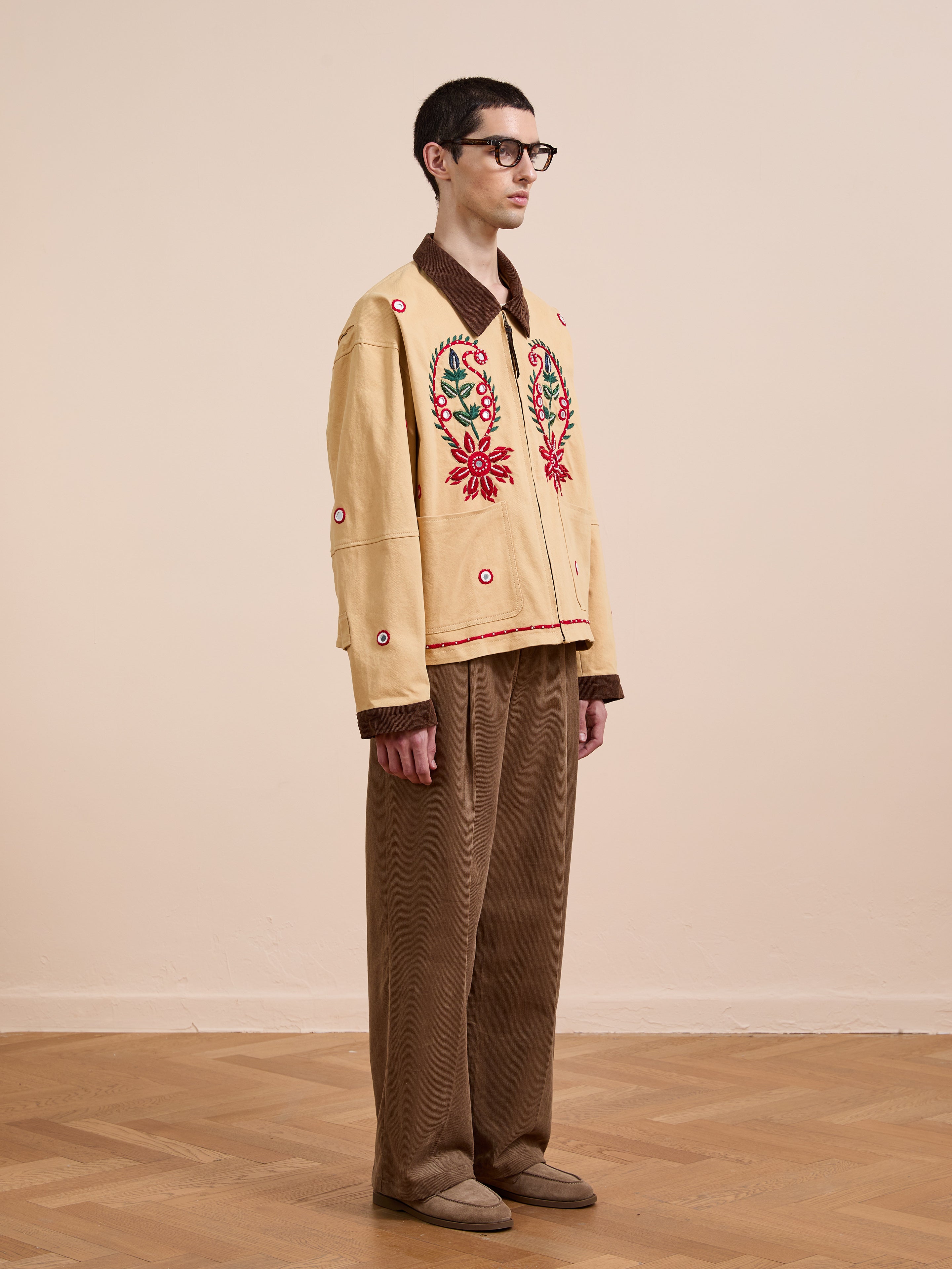 Indoors, a person wears glasses, the Found Paisley Motif Hand Embroidered Work Jacket in beige with red and green detailing, paired with brown wide-leg pants and brown shoes on a wooden floor.
