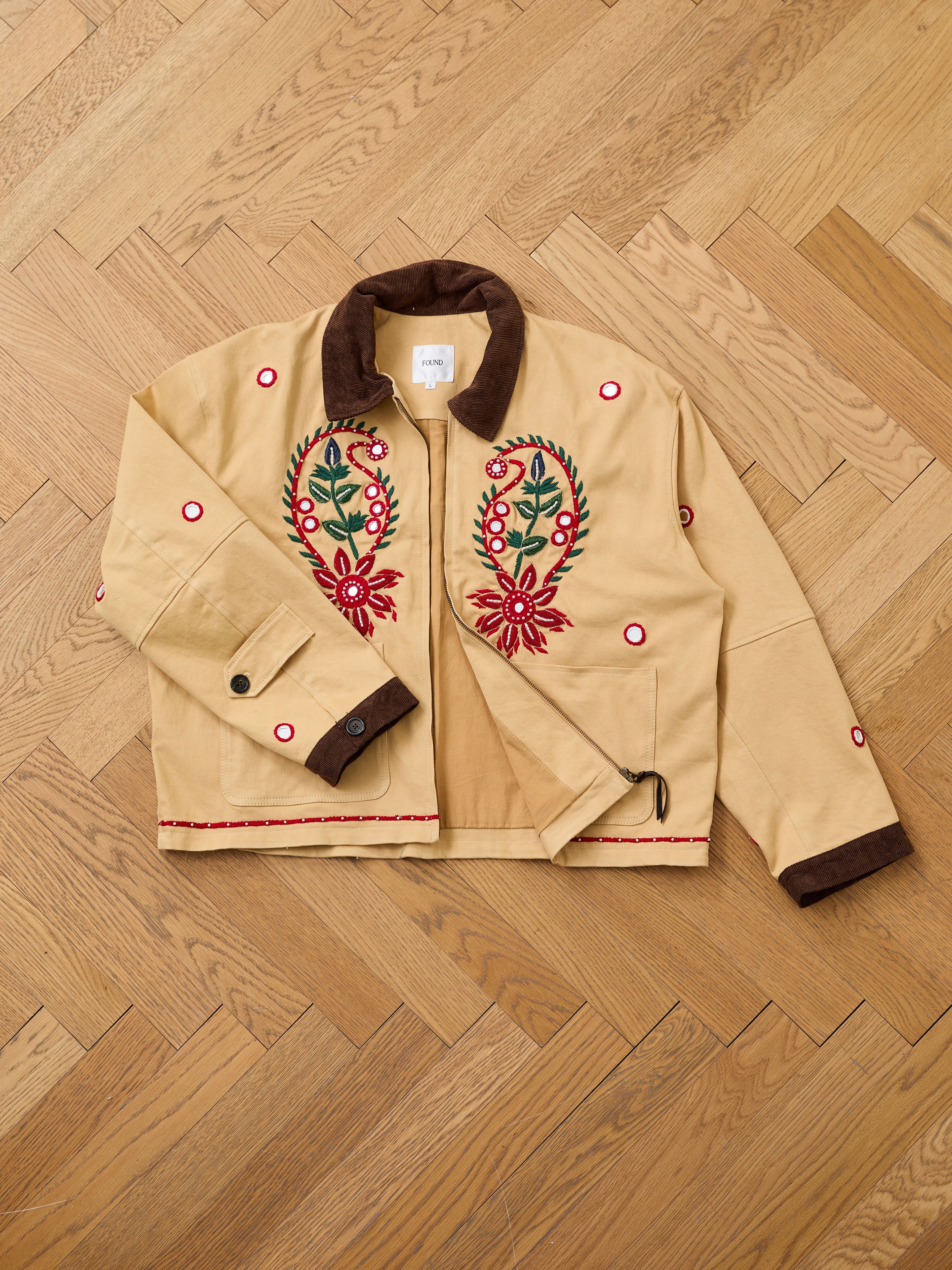 The Found Paisley Motif Hand Embroidered Work Jacket features beige fabric, a brown collar, paisley floral and circular embroidery on the front, red hem stitching, and is shown on a wooden herringbone floor.