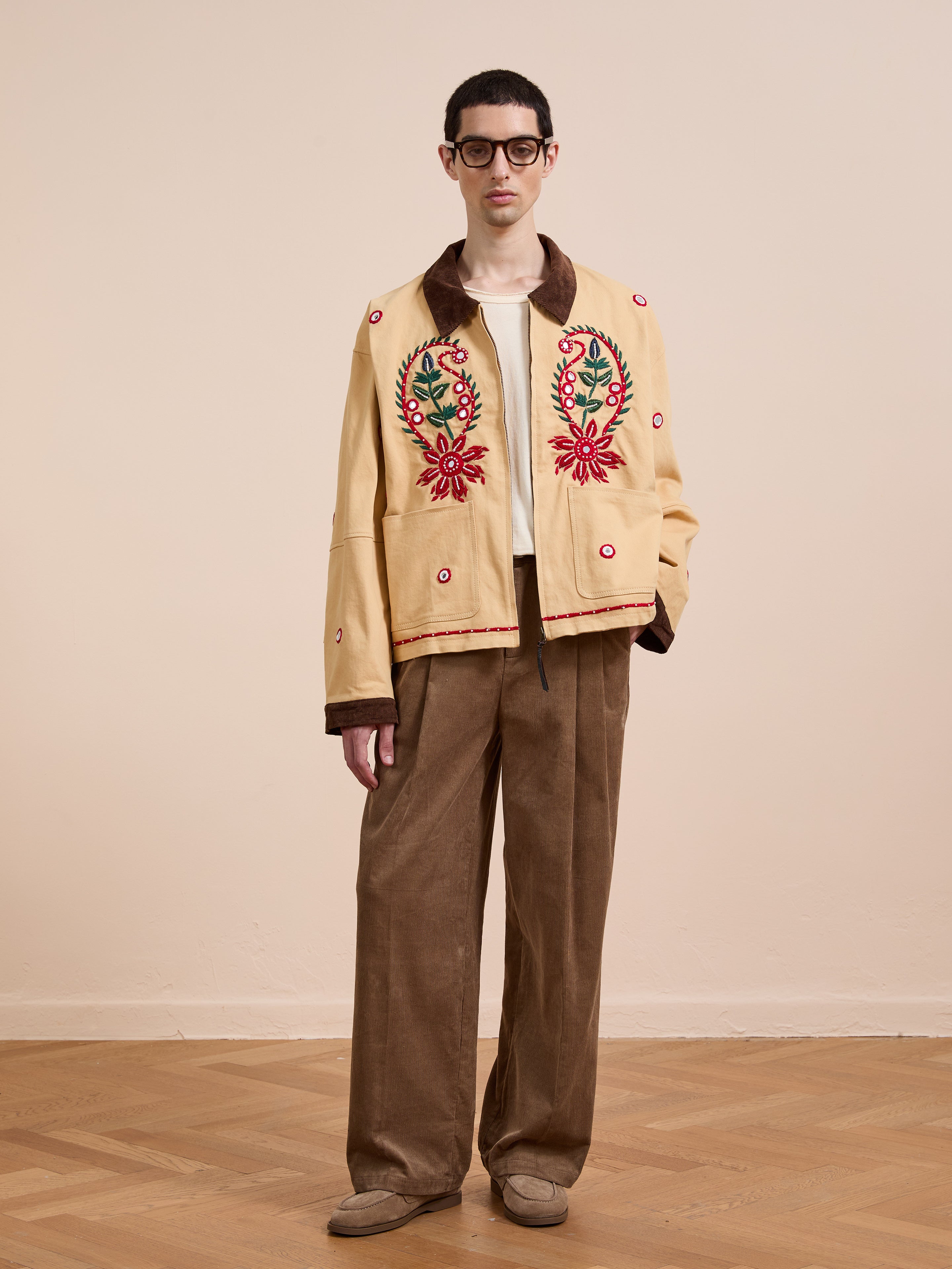 A man in glasses stands indoors wearing Found's Paisley Motif Hand Embroidered Work Jacket, paired with a cream shirt, brown wide-leg corduroy pants, and beige shoes.