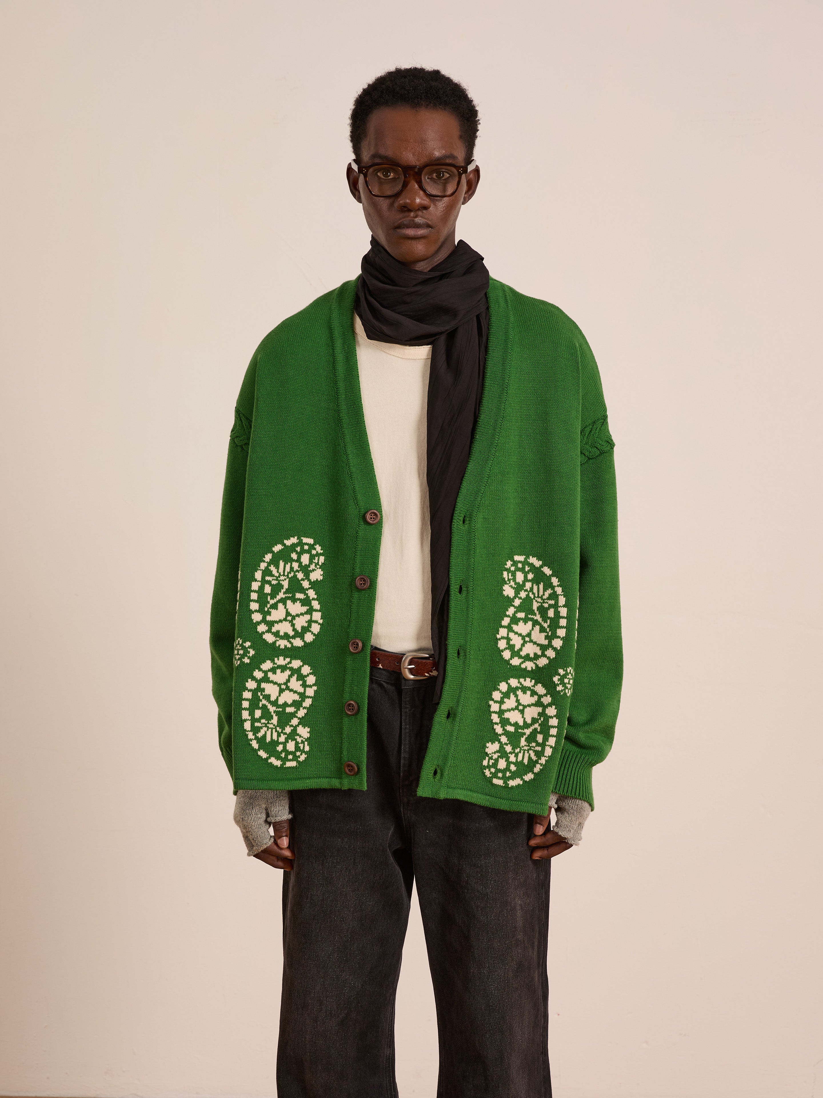 A person wearing the FOUND Paisley Knit Cardigan, featuring green cotton knit with white patterns, along with a cream shirt, dark pants, fingerless gloves, a black scarf, and glasses stands against a plain background.