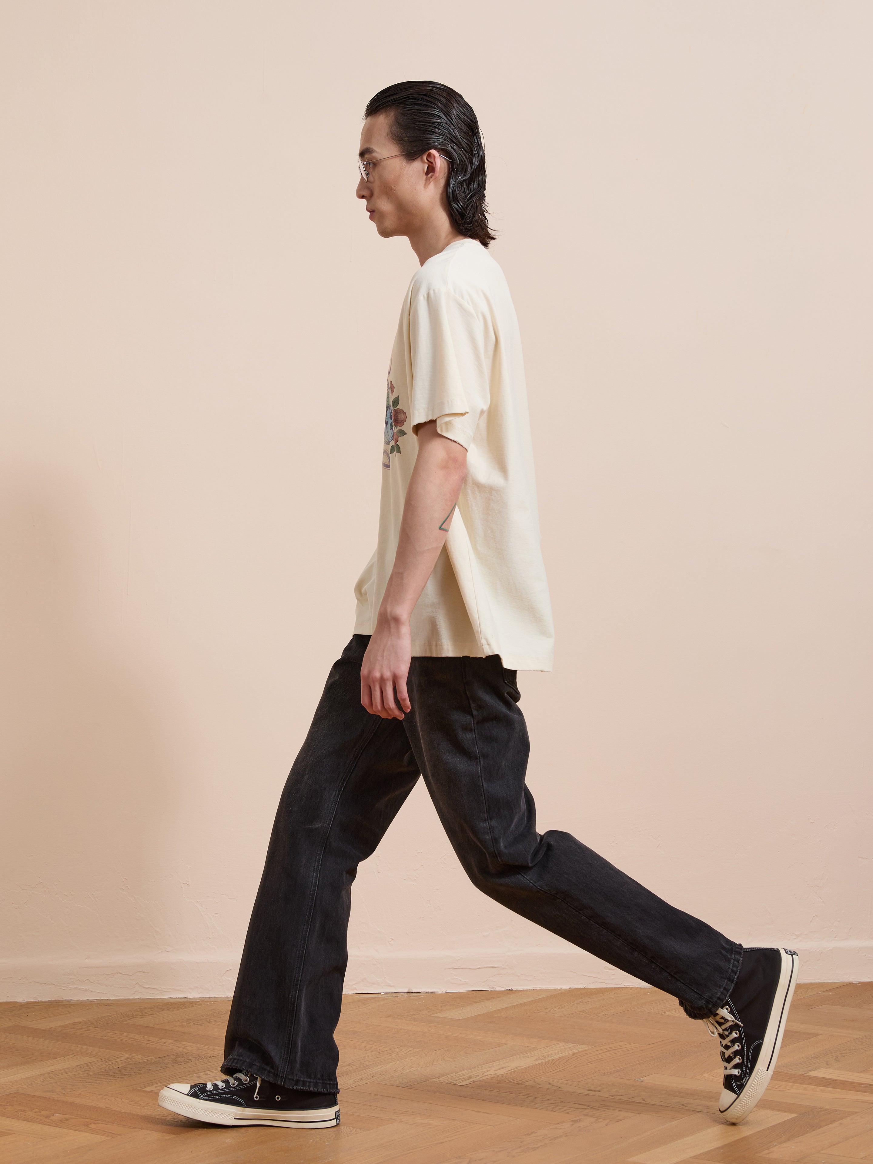 A person with long dark hair and glasses walks indoors on a wooden floor against a beige wall, wearing the FOUND Peace Globe Tee in cream vintage cotton, black jeans, and black sneakers.