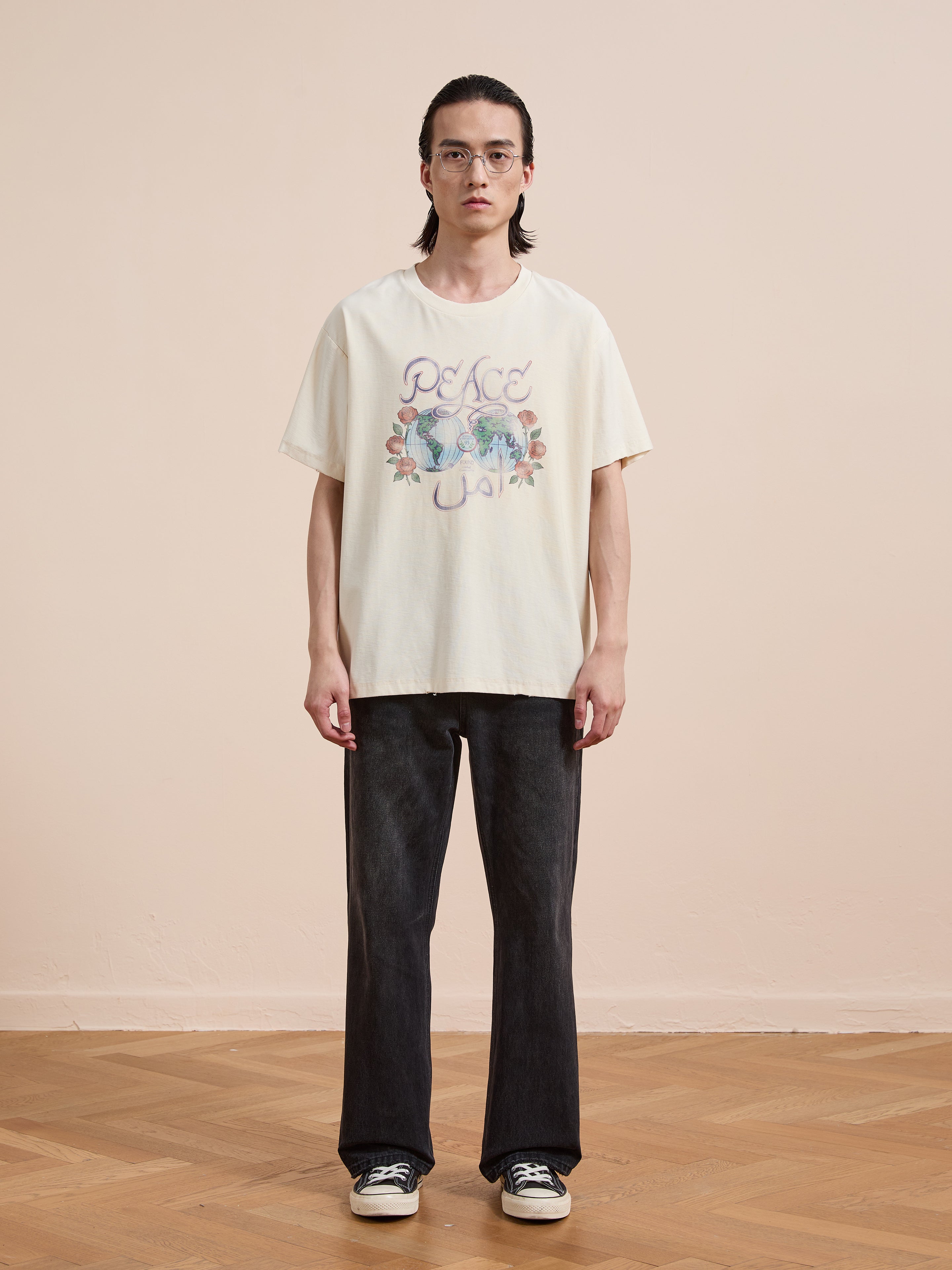 Indoors on a wooden floor, a person in glasses, black jeans, and sneakers wears the FOUND Peace Globe Tee—a cream-colored vintage cotton shirt with “Peace and Love” graphics—against a plain beige wall.
