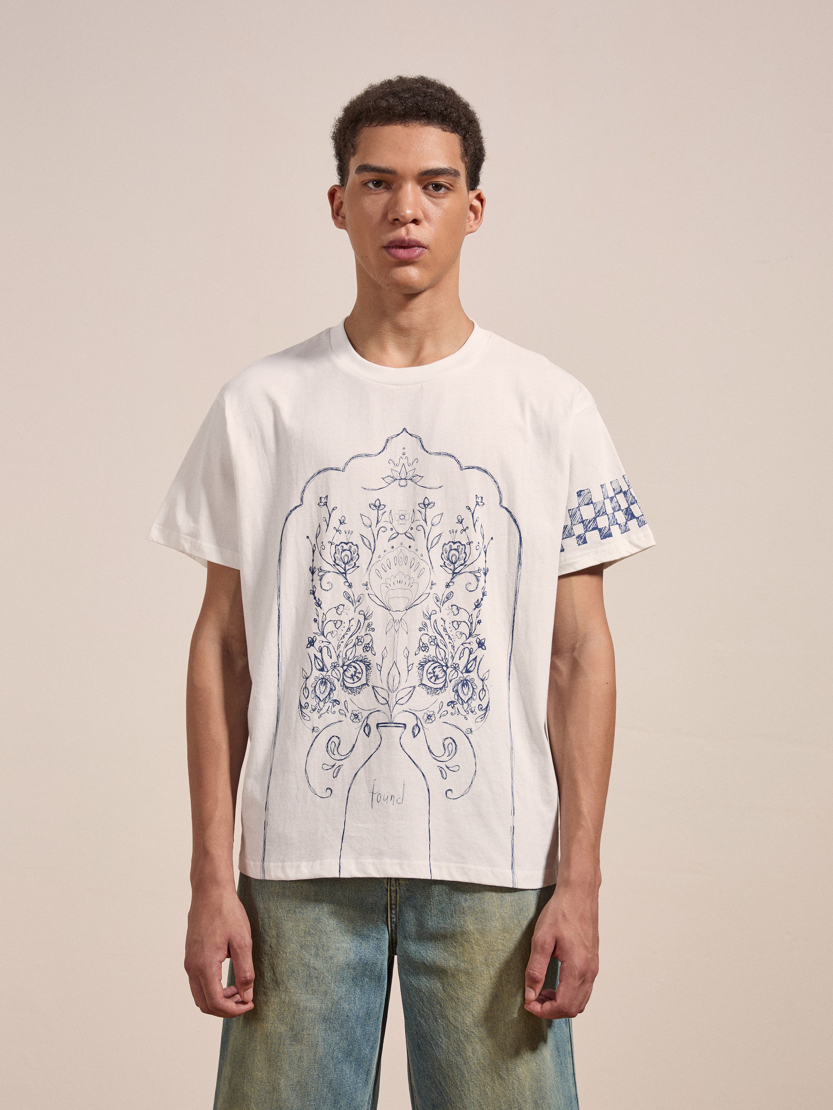 A person stands wearing the "Pen Flower Medley" white T-shirt by FOUND, featuring a blue ornamental floral design on the front and patterned details on the sleeves. They are also wearing light blue jeans.