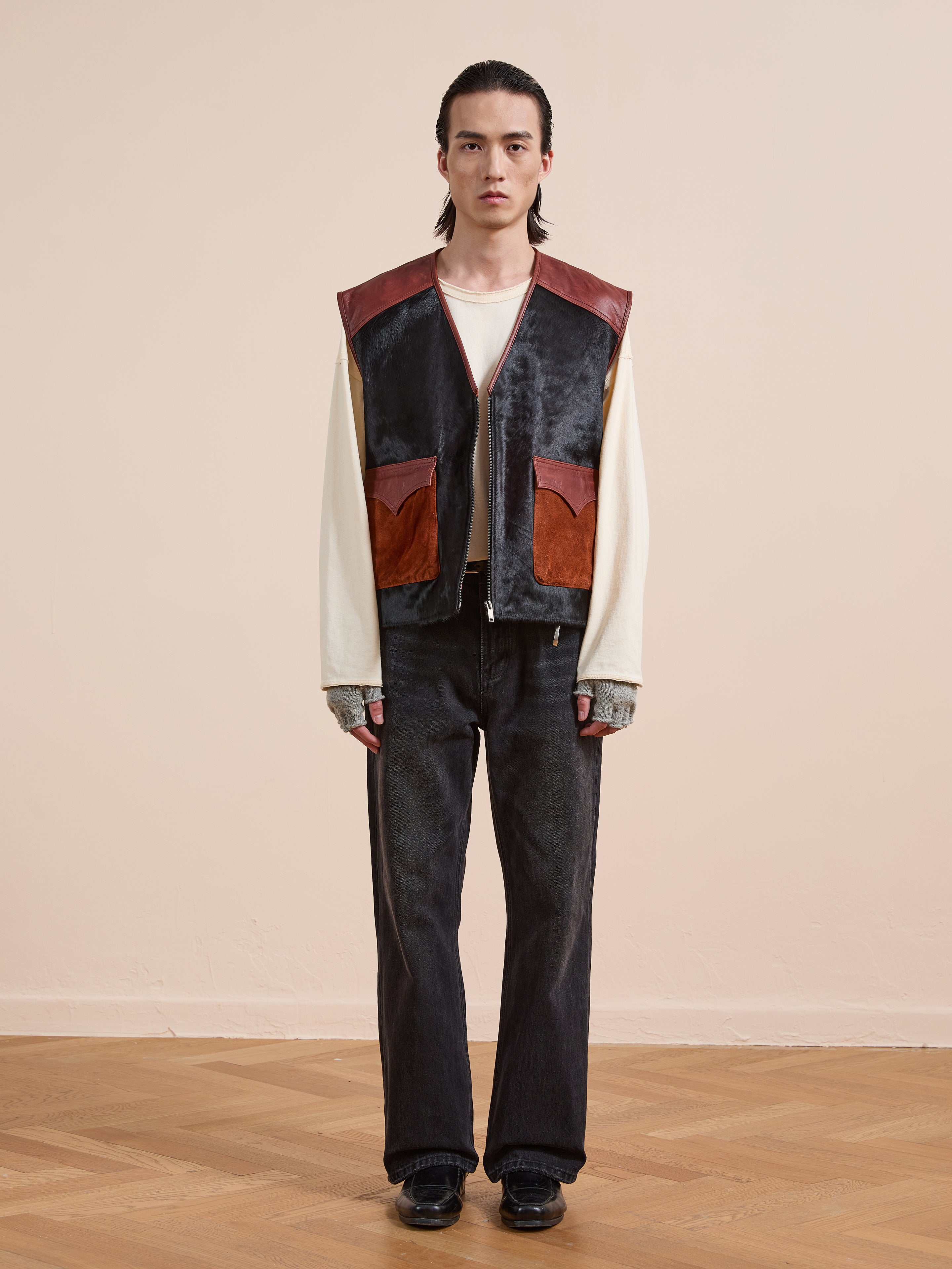 A person stands indoors against a beige wall wearing Found’s Fur Western Leather Vest in two-tone brown and black, paired with a cream long-sleeve shirt, black jeans, gray gloves, and black shoes.