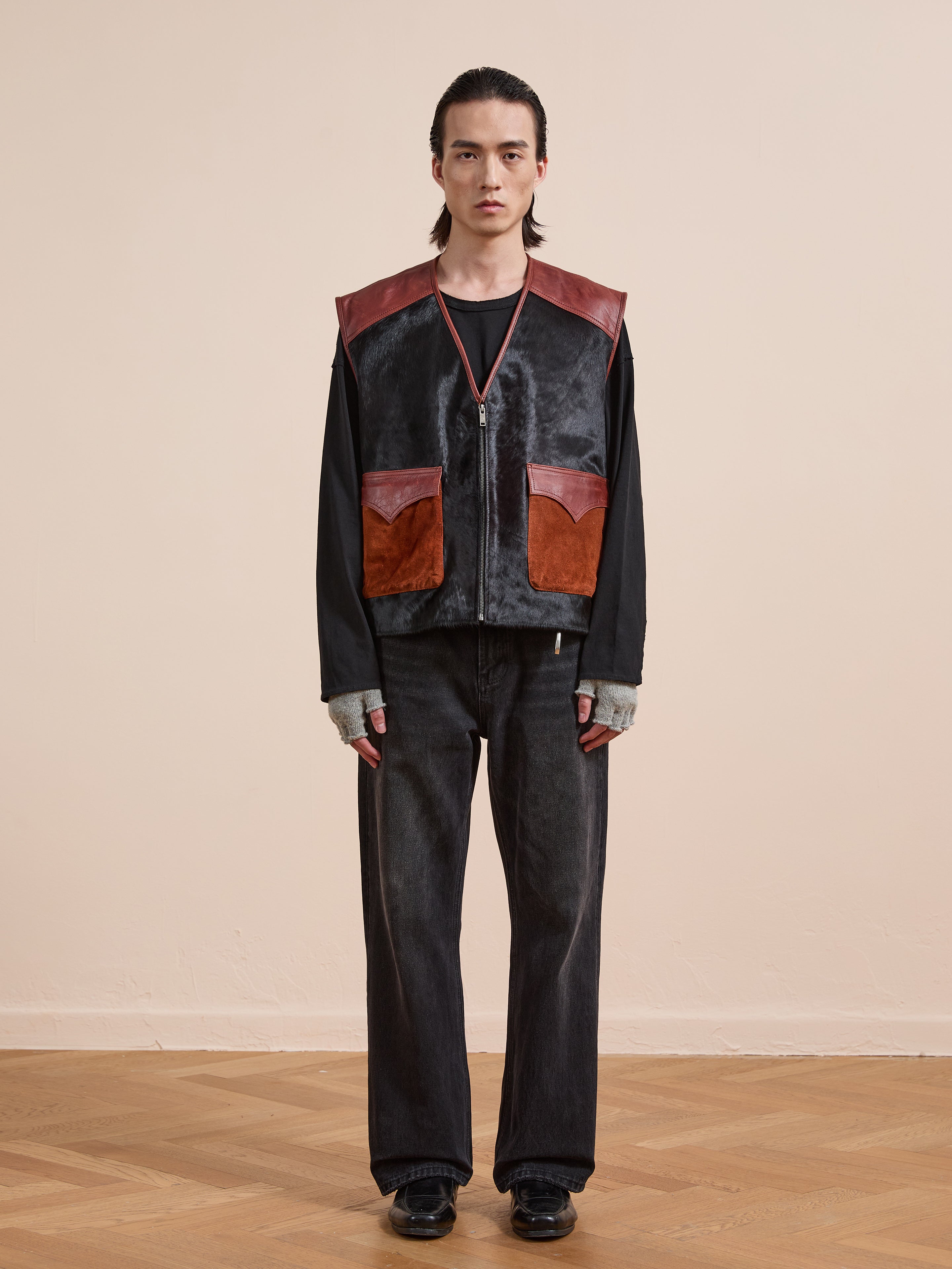 A person stands indoors against a plain beige wall wearing the Found Fur Western Leather Vest in black and brown over a black shirt, paired with dark pants, fingerless gloves, and black shoes.