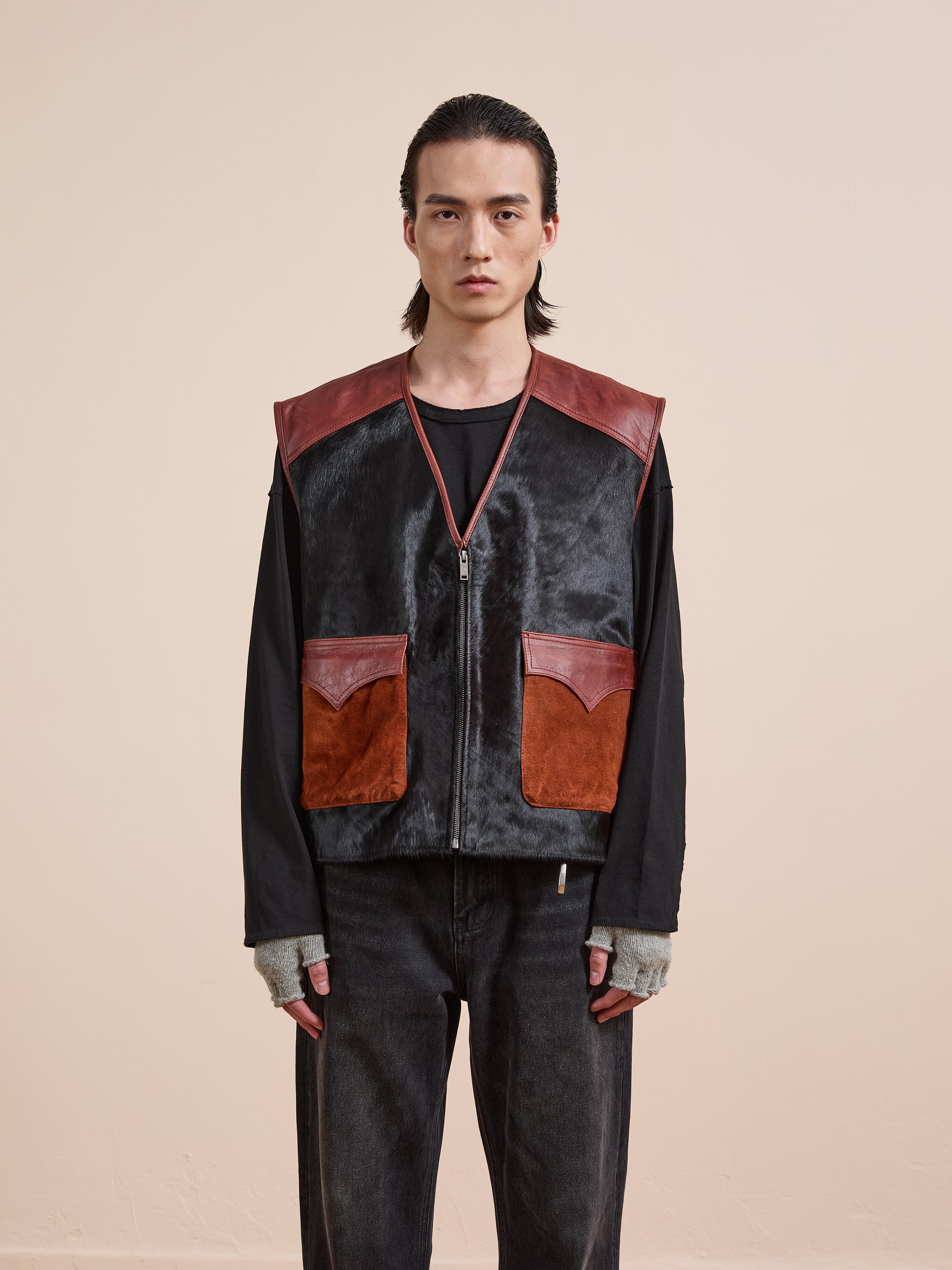 A person stands facing forward in a black long-sleeve shirt, black pants, and the Found Fur Western Leather Vest featuring brown leather accents and front pockets, set against a plain beige background.