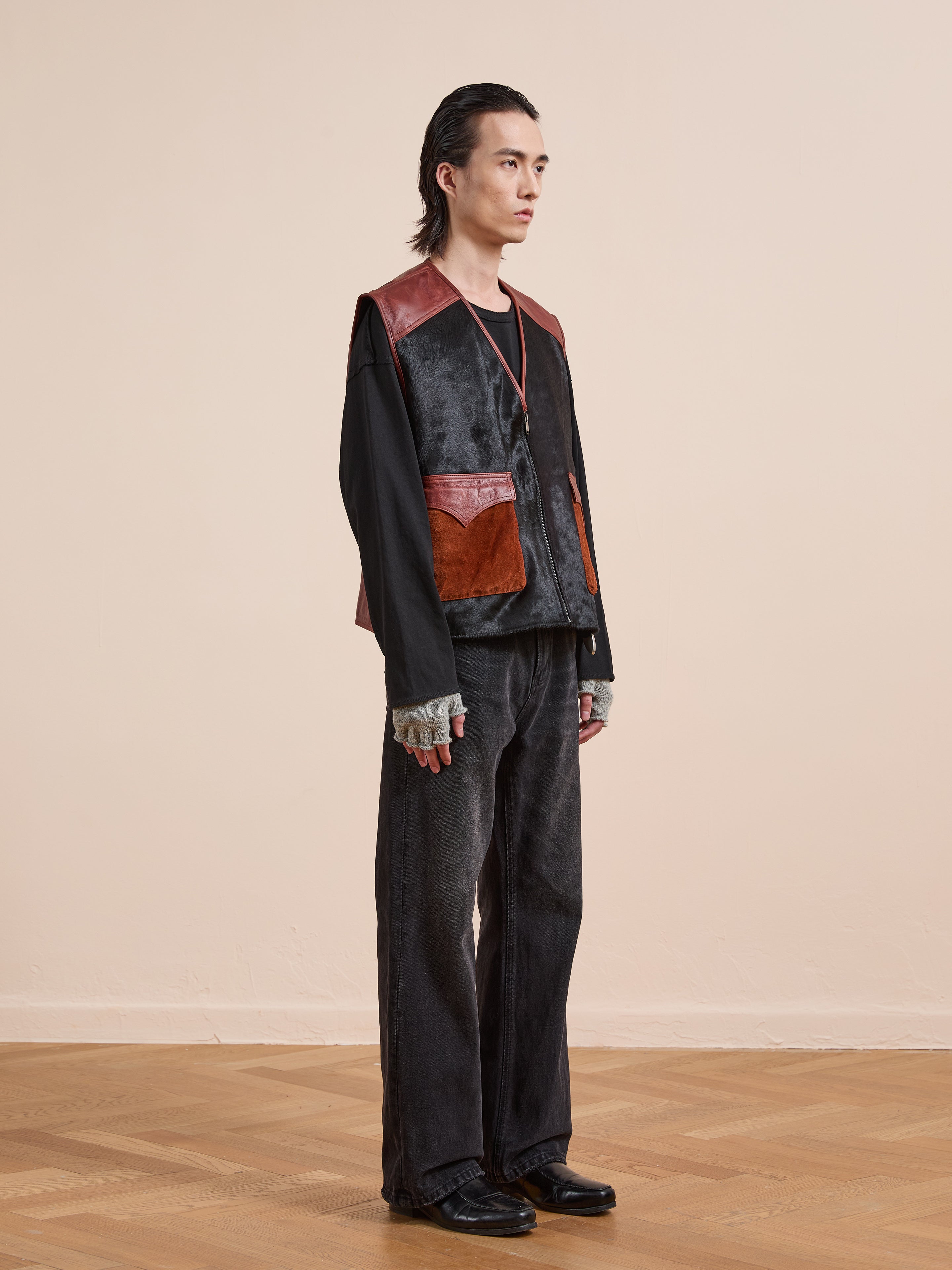 A person stands indoors wearing a black long-sleeve shirt, black wide-leg pants, Found's Fur Western Leather Vest, fingerless gloves, and black shoes.