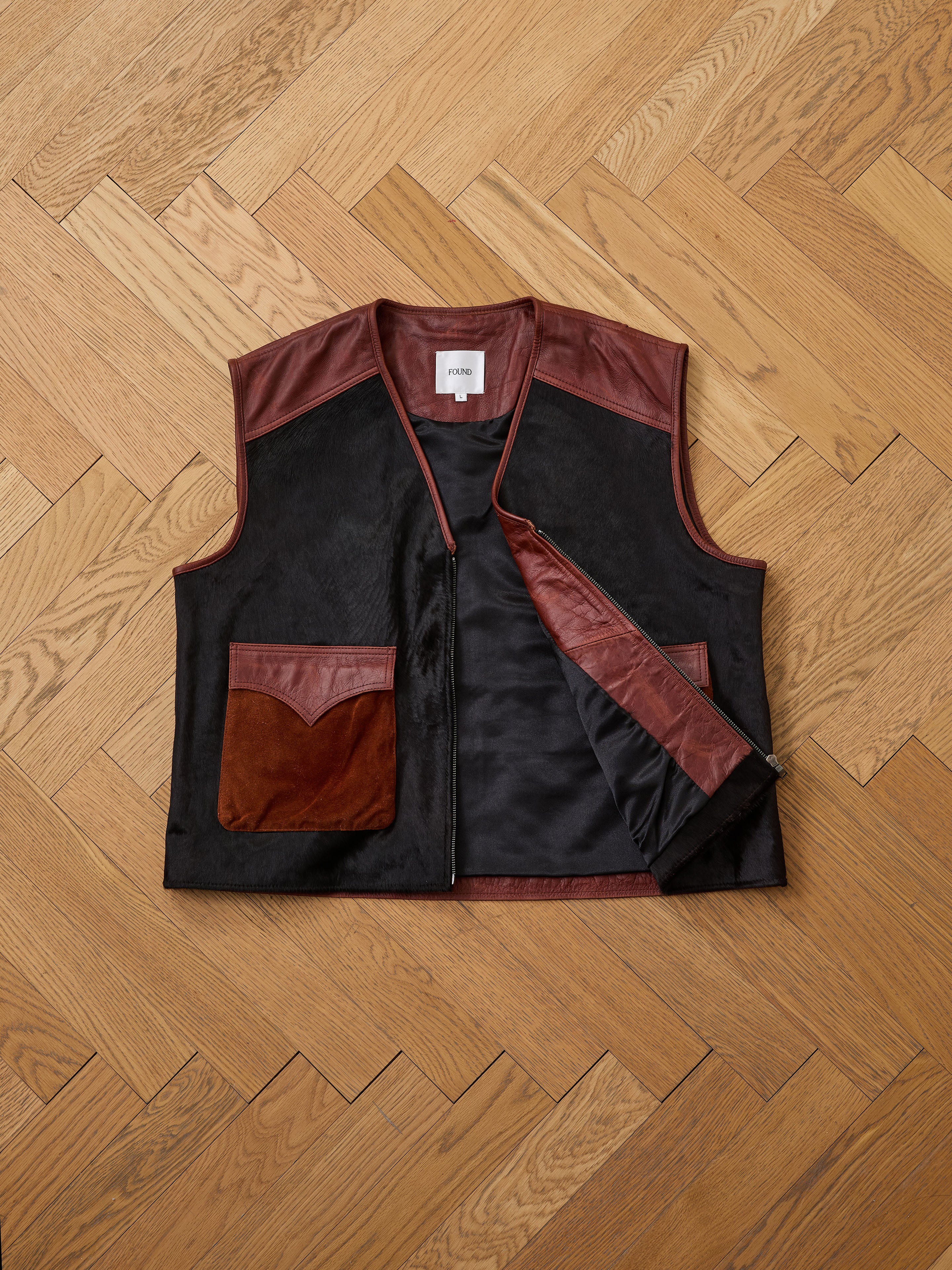 The Found Fur Western Leather Vest is a sleeveless black vest with brown leather accents, two front pockets, and a front zipper, shown laid flat on a wooden herringbone floor.