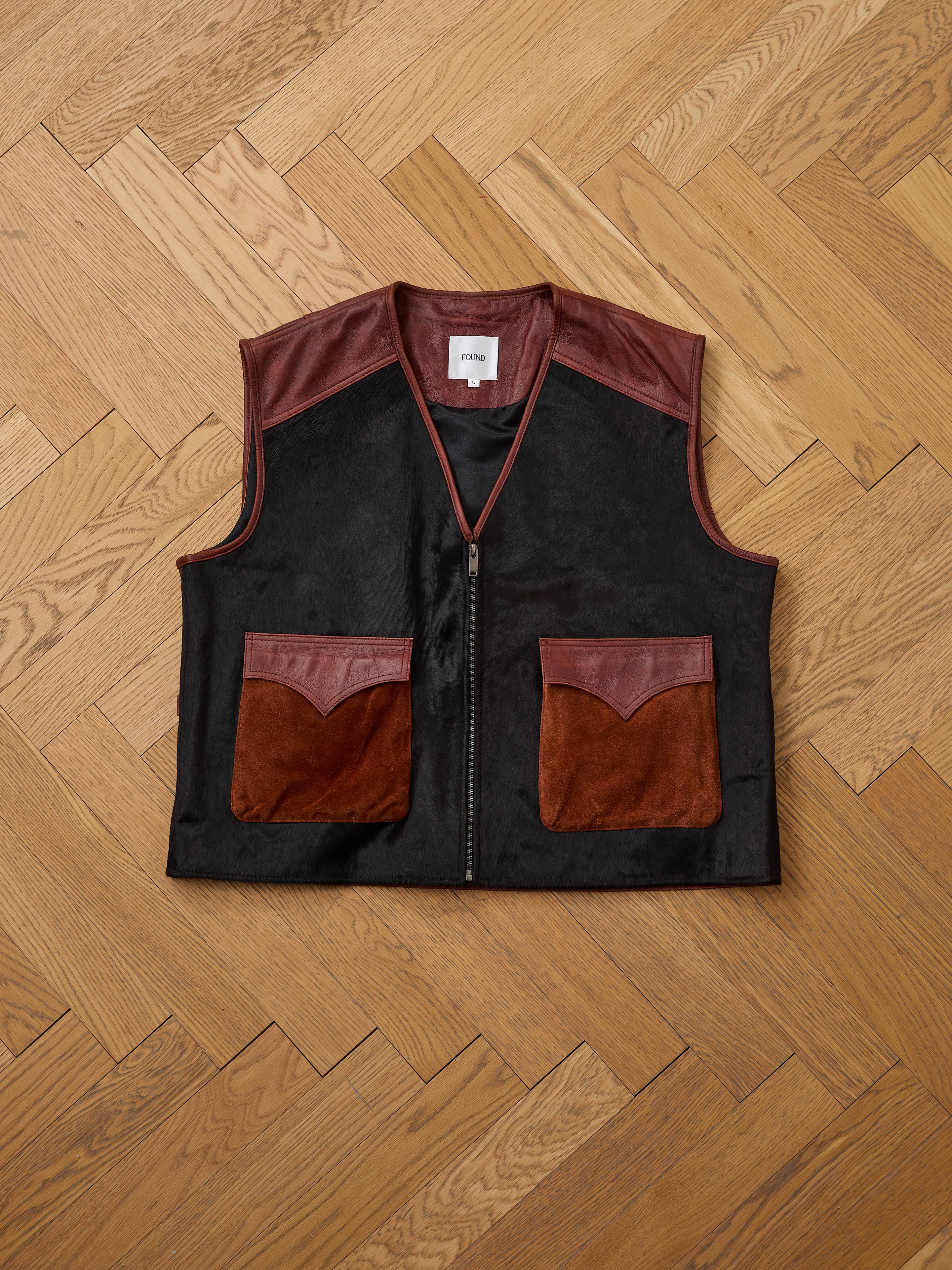 The Found Fur Western Leather Vest is a sleeveless black vest with brown leather shoulder accents and two front pockets, shown on a wooden herringbone floor.