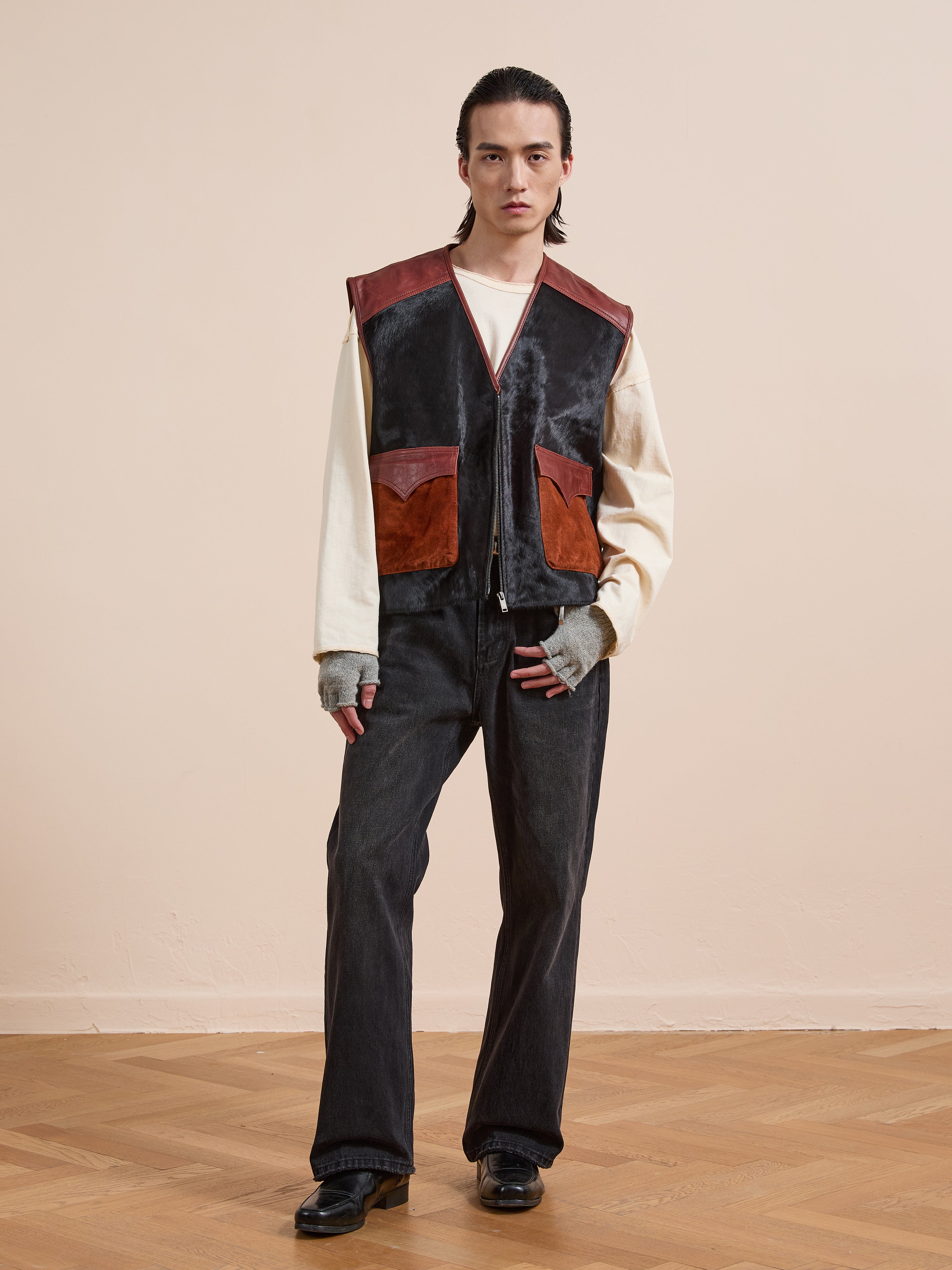 A person stands indoors on a wooden floor, wearing a cream shirt, dark jeans, black shoes, and the Found Fur Western Leather Vest in black and brown, looking directly at the camera.