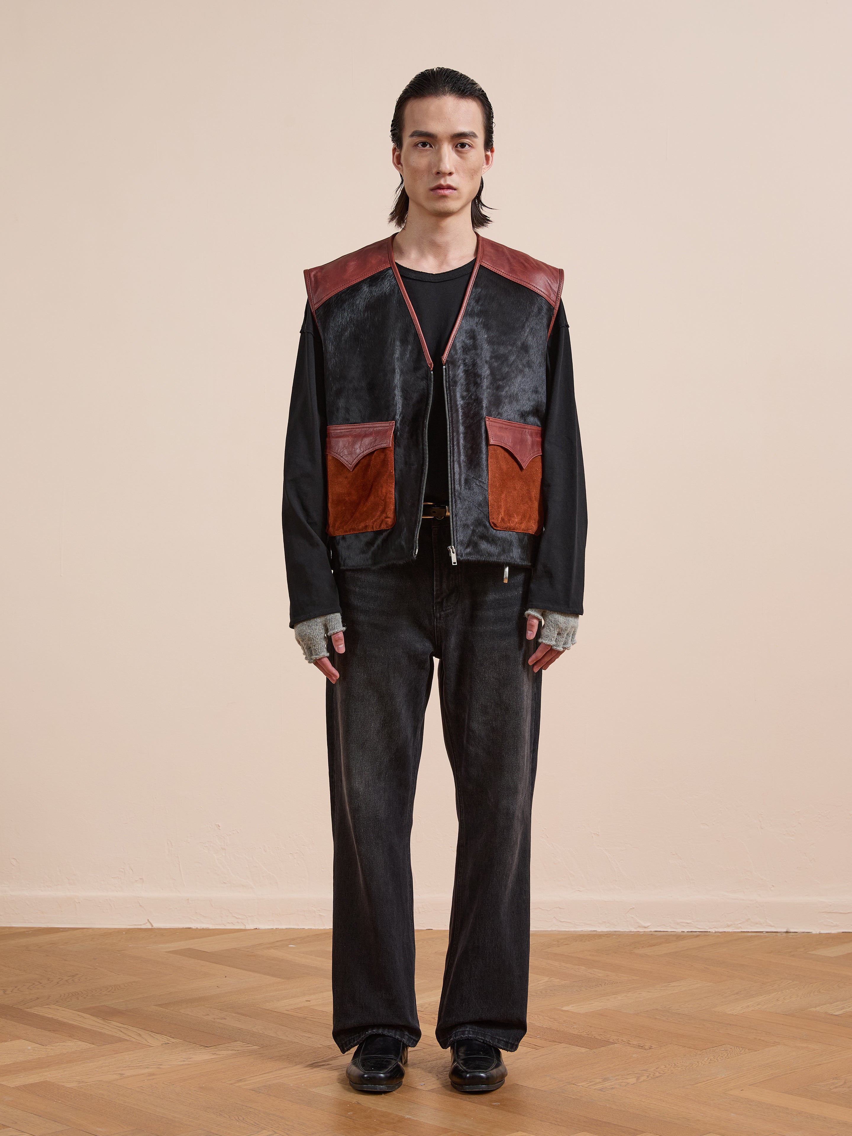 A person stands facing forward, wearing a black shirt, black jeans, gray fingerless gloves, black shoes, and the Found Fur Western Leather Vest with large black and brown pockets, against a plain beige wall.