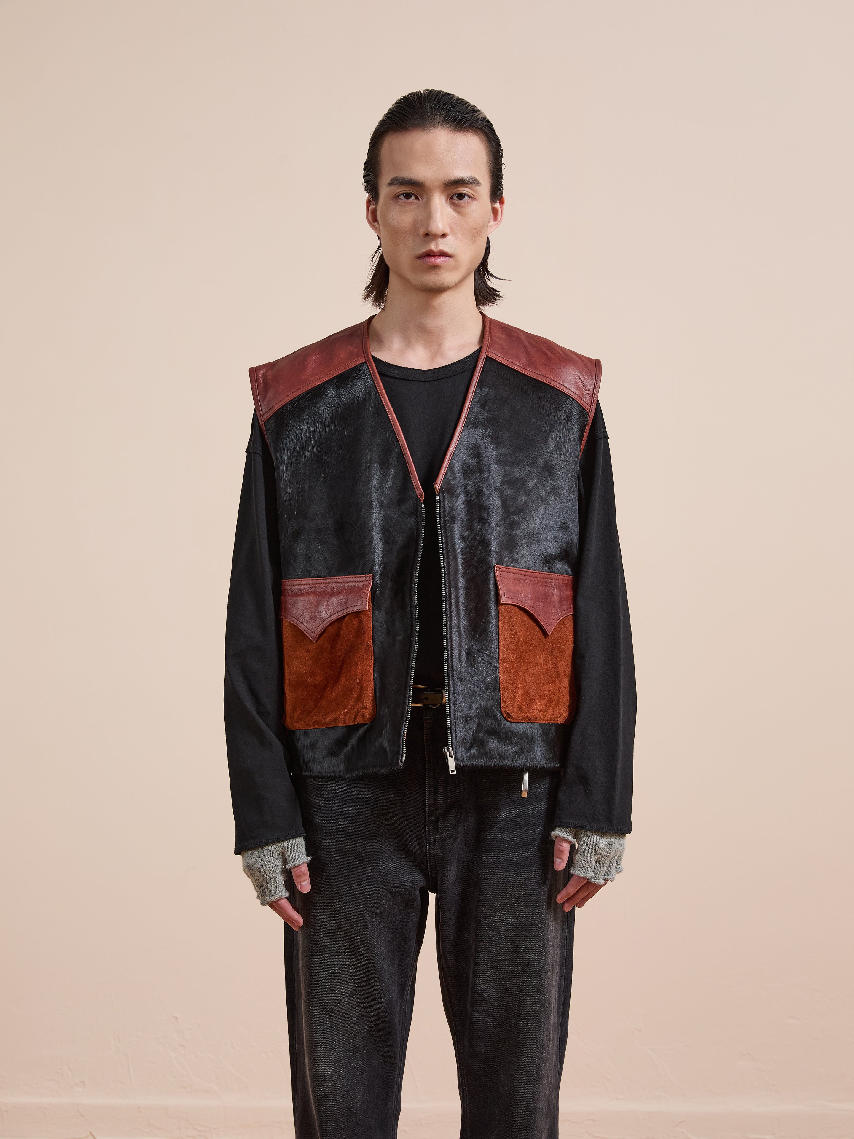 A person stands facing forward against a plain beige background, wearing a black shirt, dark jeans, fingerless gray gloves, and the Found Fur Western Leather Vest featuring large black and brown pockets.