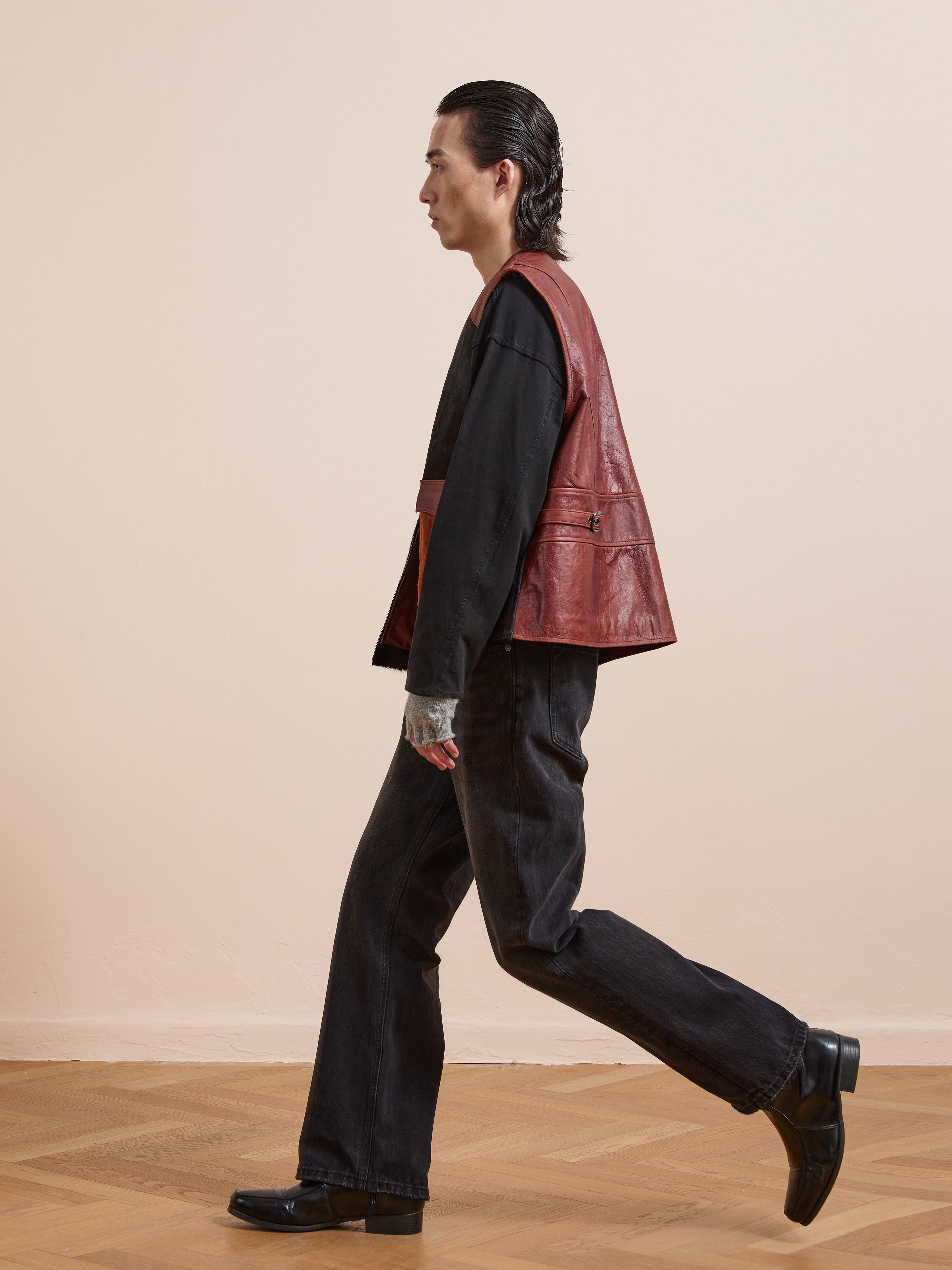 A person with slicked-back hair walks indoors, wearing Found’s Fur Western Leather Vest in red over a black shirt, black jeans, gray gloves, and black shoes.