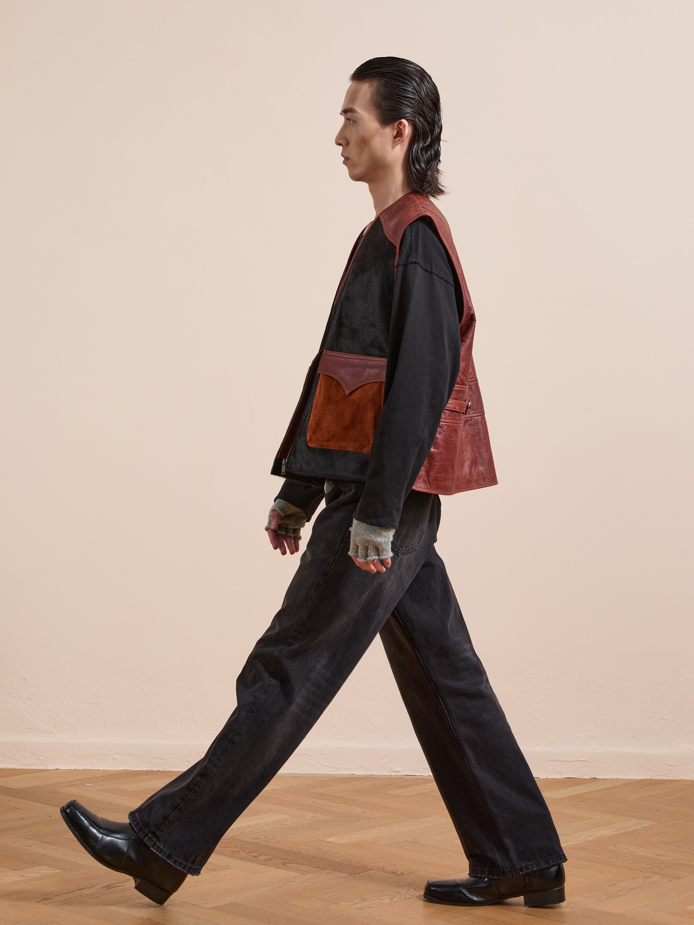 A person walks indoors in a black long-sleeve shirt, wide black jeans, and black shoes, styled with the Found Fur Western Leather Vest—a brown leather vest with large pockets—set against a plain beige wall.