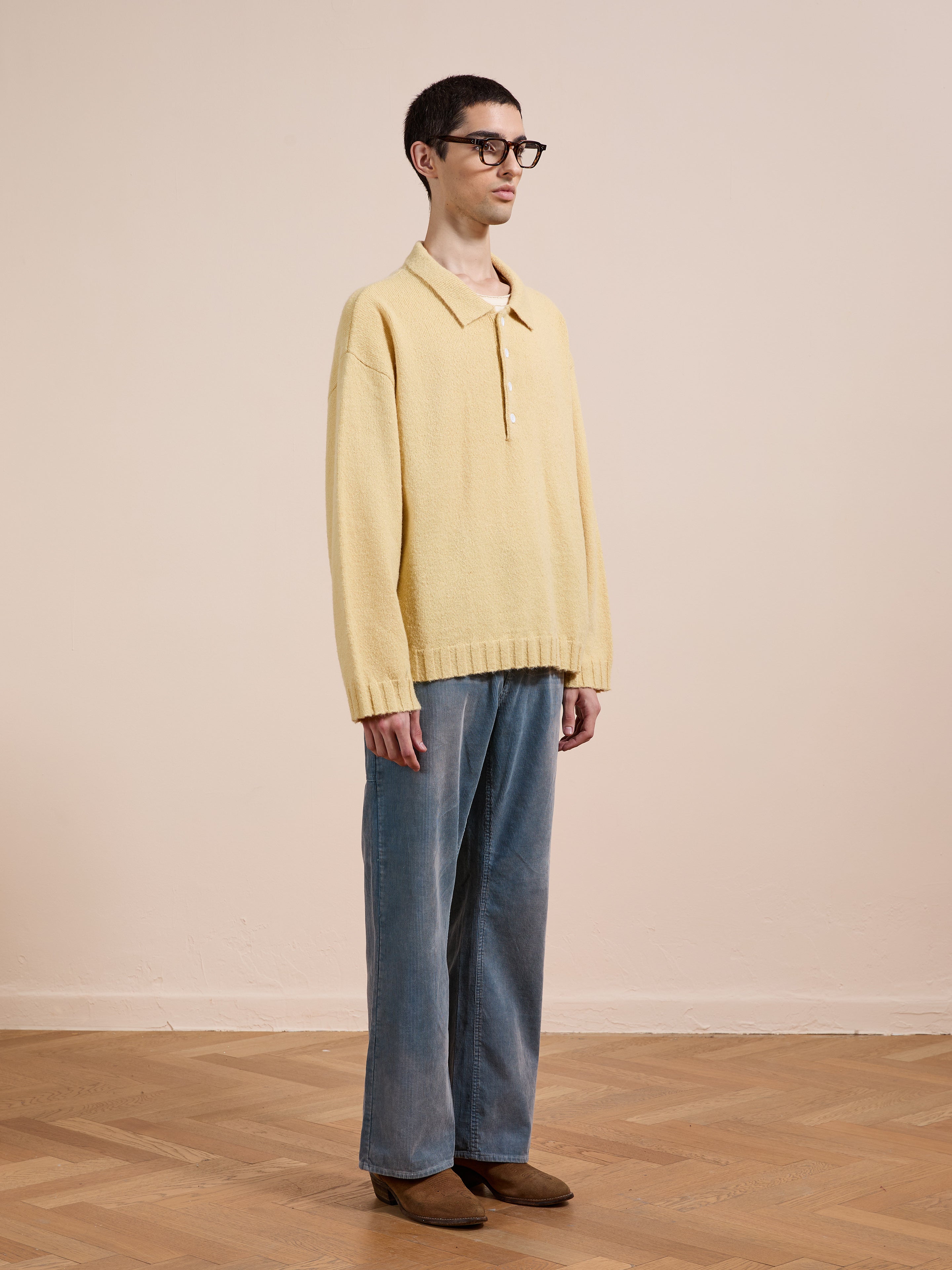 A person with short dark hair and glasses stands indoors against a beige background, wearing the FOUND Knit LS Polo Sweater and light blue jeans with brown shoes.