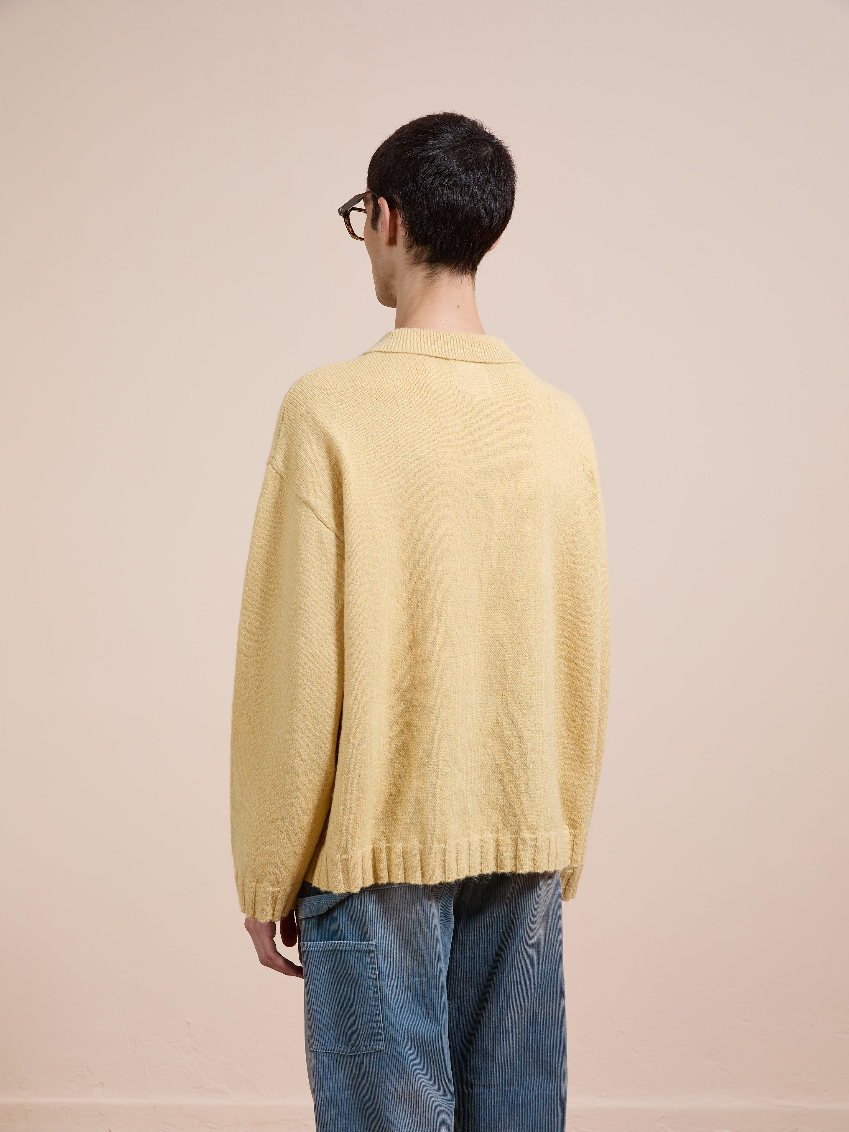 A person with short dark hair and glasses, seen from behind, wears the FOUND Knit LS Polo Sweater—a loose unisex style in brushed knit—paired with blue jeans against a plain light background.