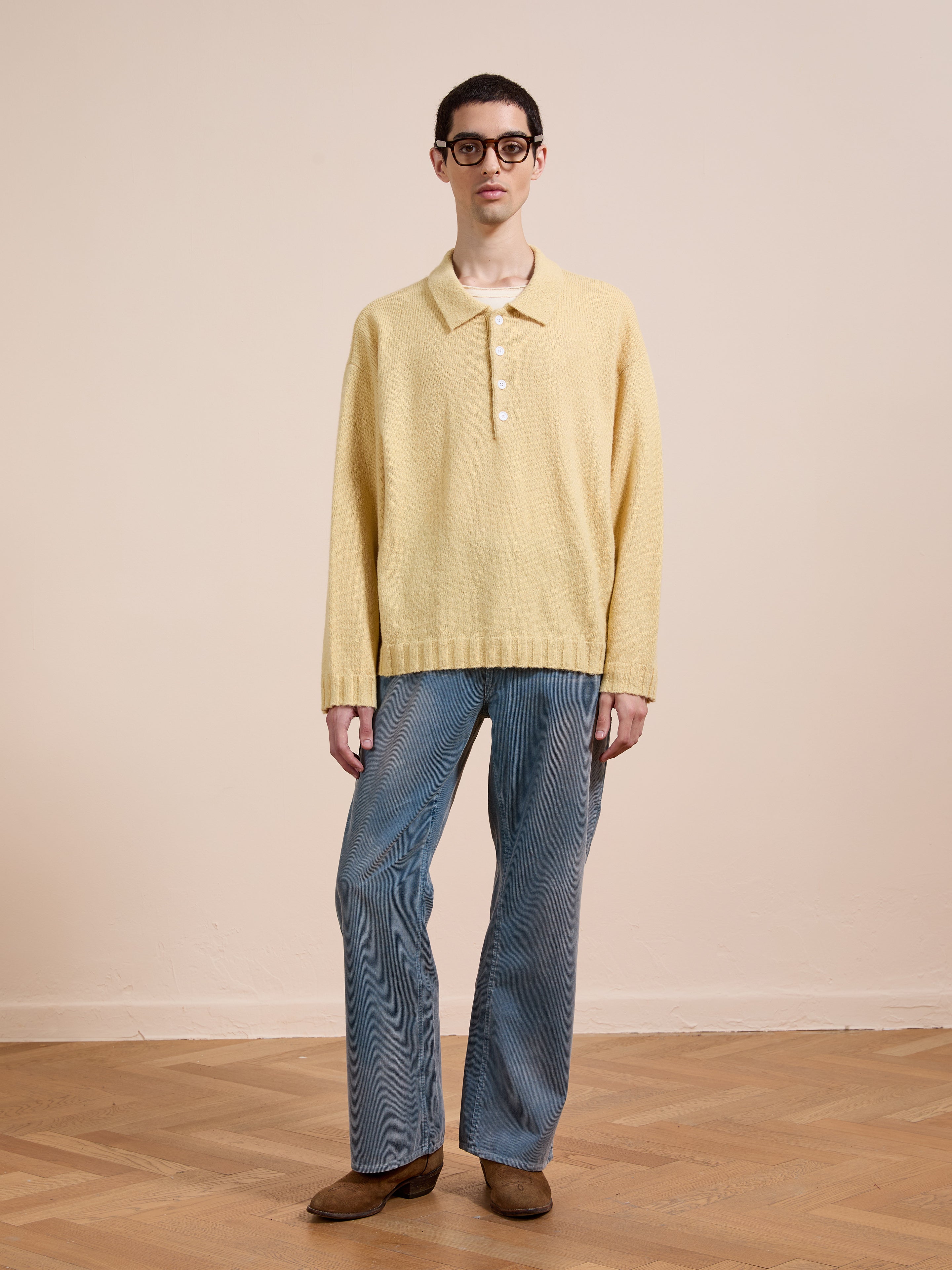 A person wearing the FOUND Knit LS Polo Sweater in yellow, blue jeans, brown shoes, and glasses stands on a wooden floor against a plain beige background.