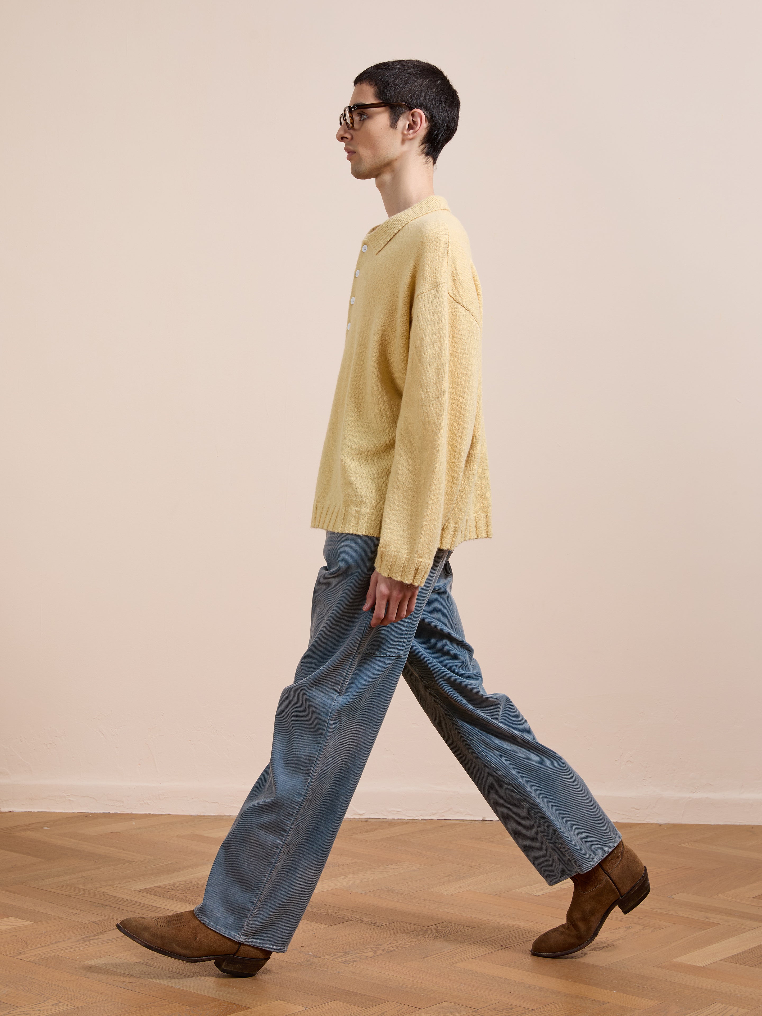 A person in the FOUND Knit LS Polo Sweater in yellow, blue wide-leg jeans, and brown shoes walks indoors on a wooden floor with a plain beige wall background.