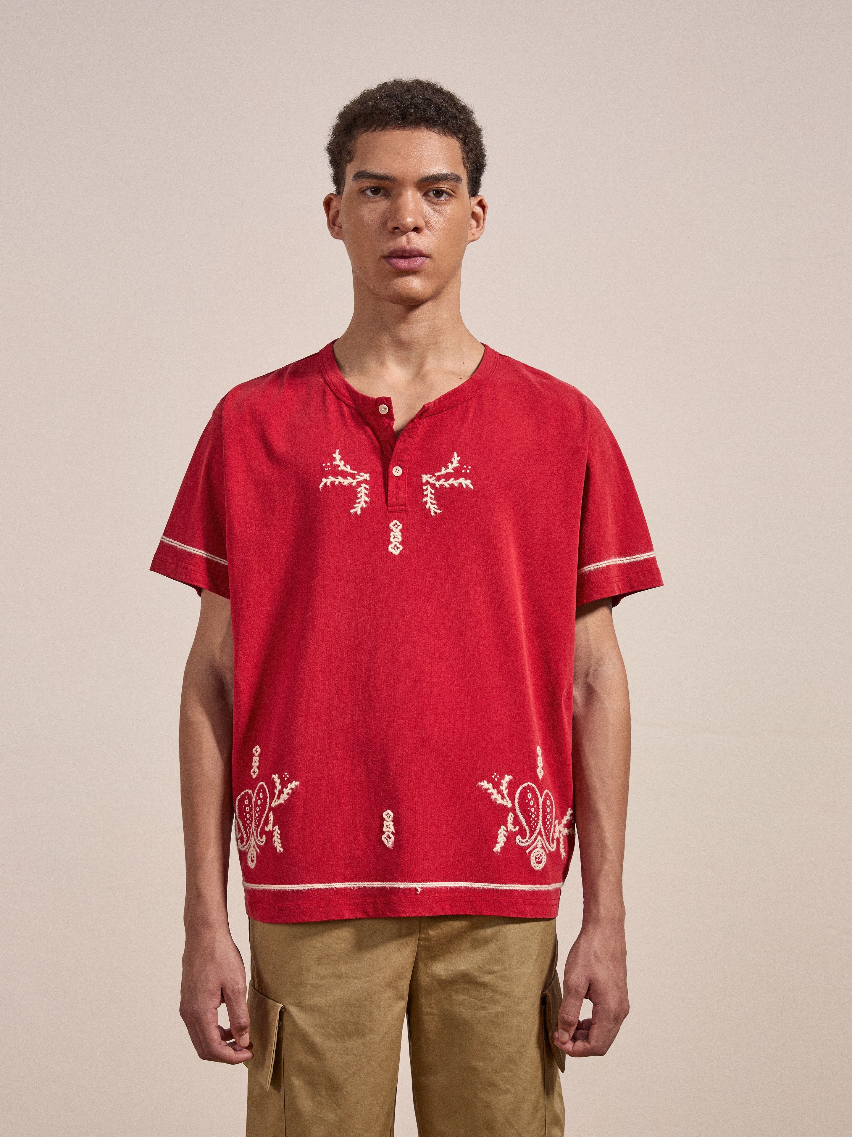 A person stands against a plain background, wearing the FOUND Embroidered Henley, a red short-sleeved shirt with white embroidered patterns, and beige pants.