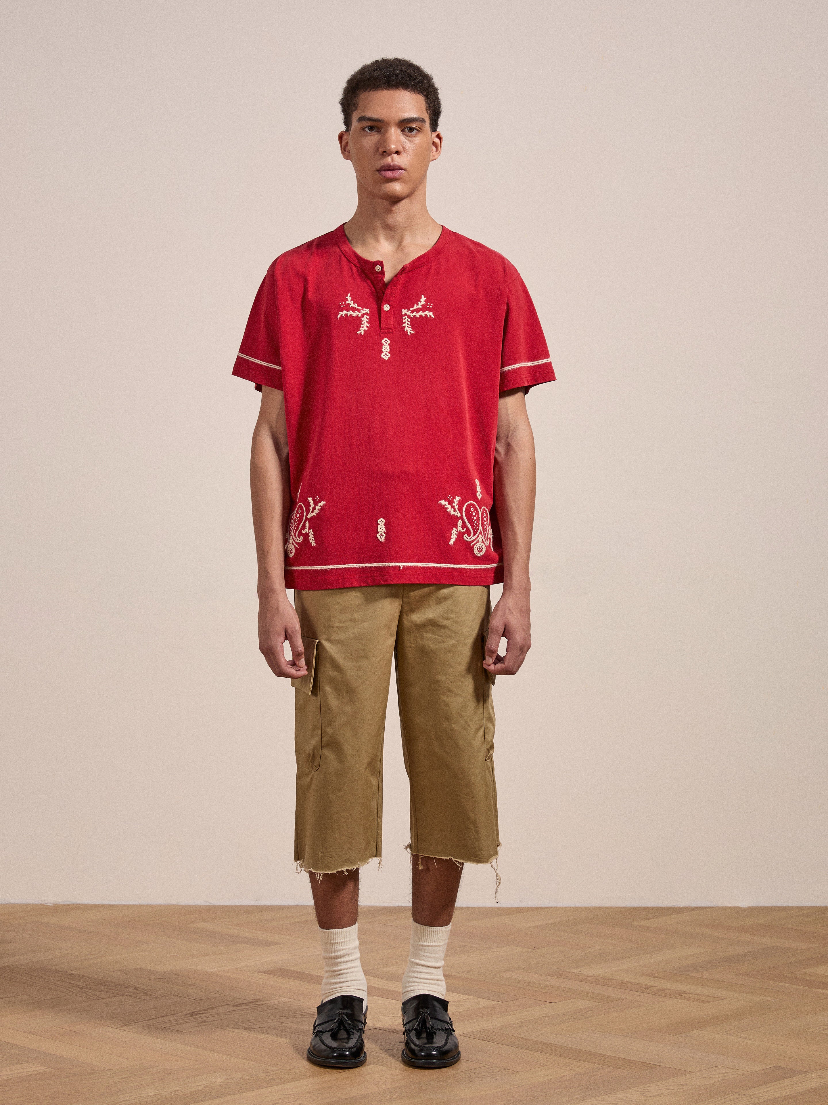 A person stands indoors wearing a red Embroidered Henley by FOUND, beige knee-length shorts, white socks, and black shoes. They are posed against a plain, neutral-colored background.