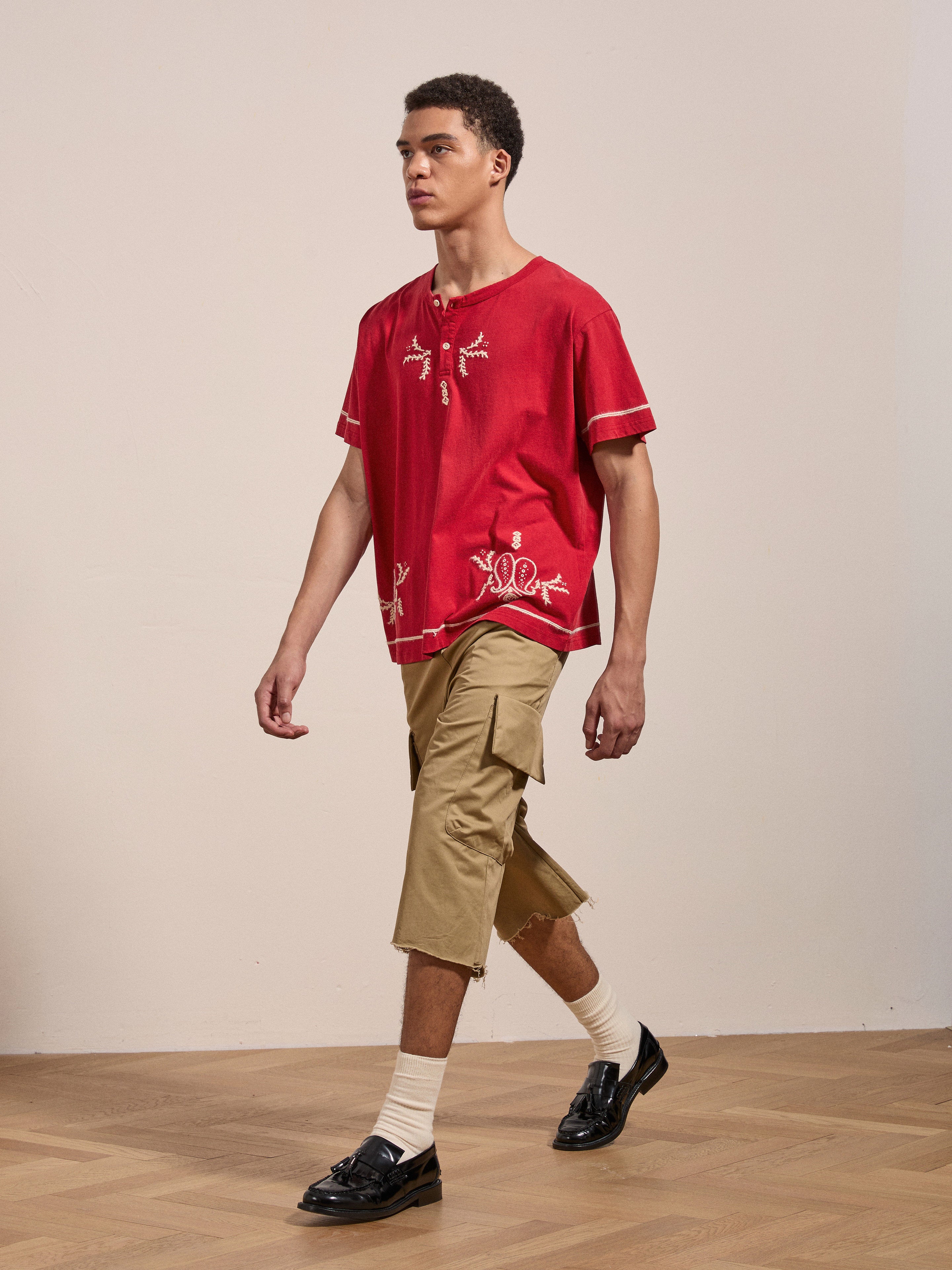 A person walks on a wooden floor, wearing the FOUND Embroidered Henley in red, beige cargo shorts, white socks, and black loafers.