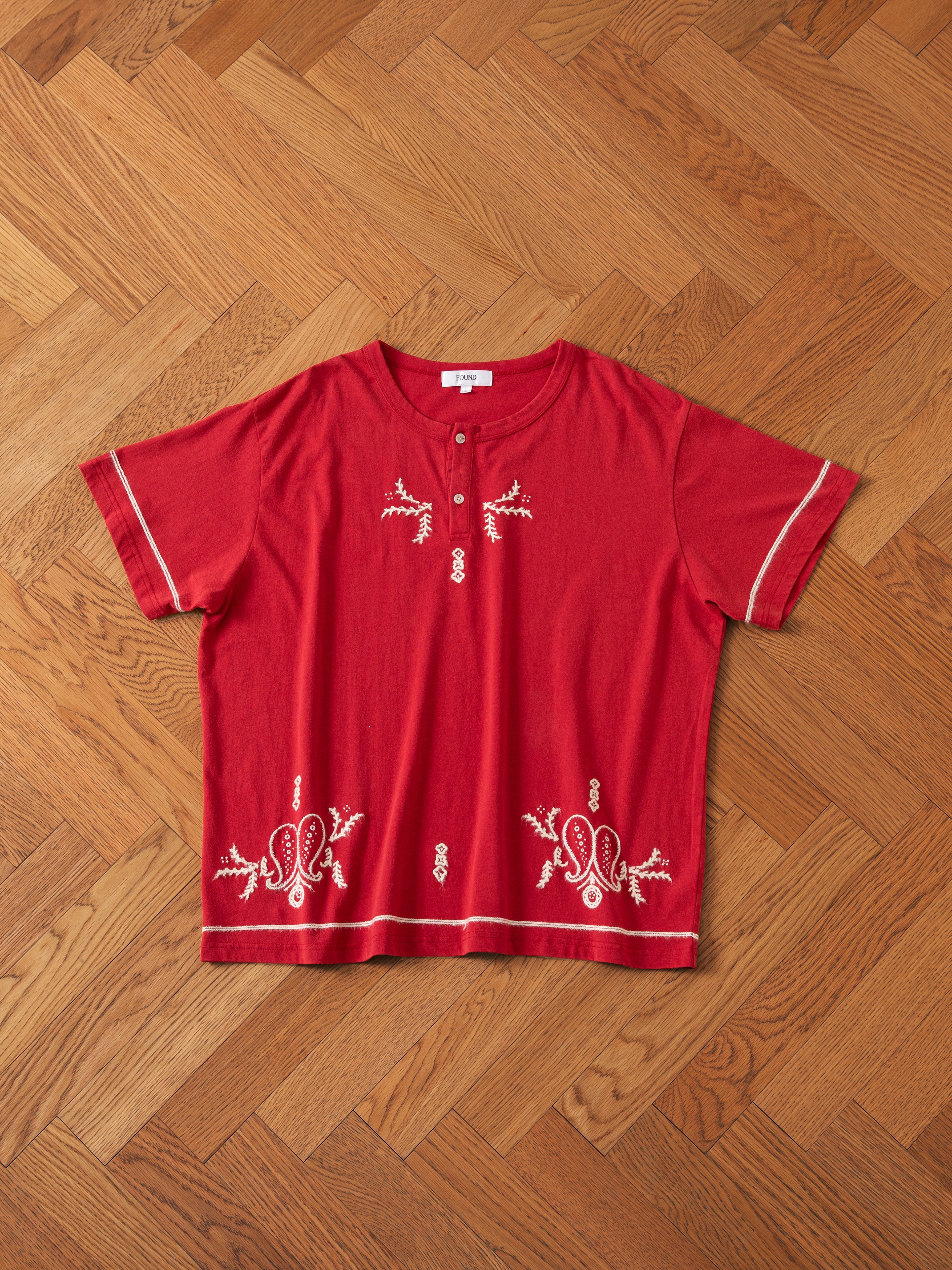 An Embroidered Henley by FOUND, featuring a red short-sleeve design with decorative white embroidery on the front and cuffs, displayed on a wooden parquet floor.