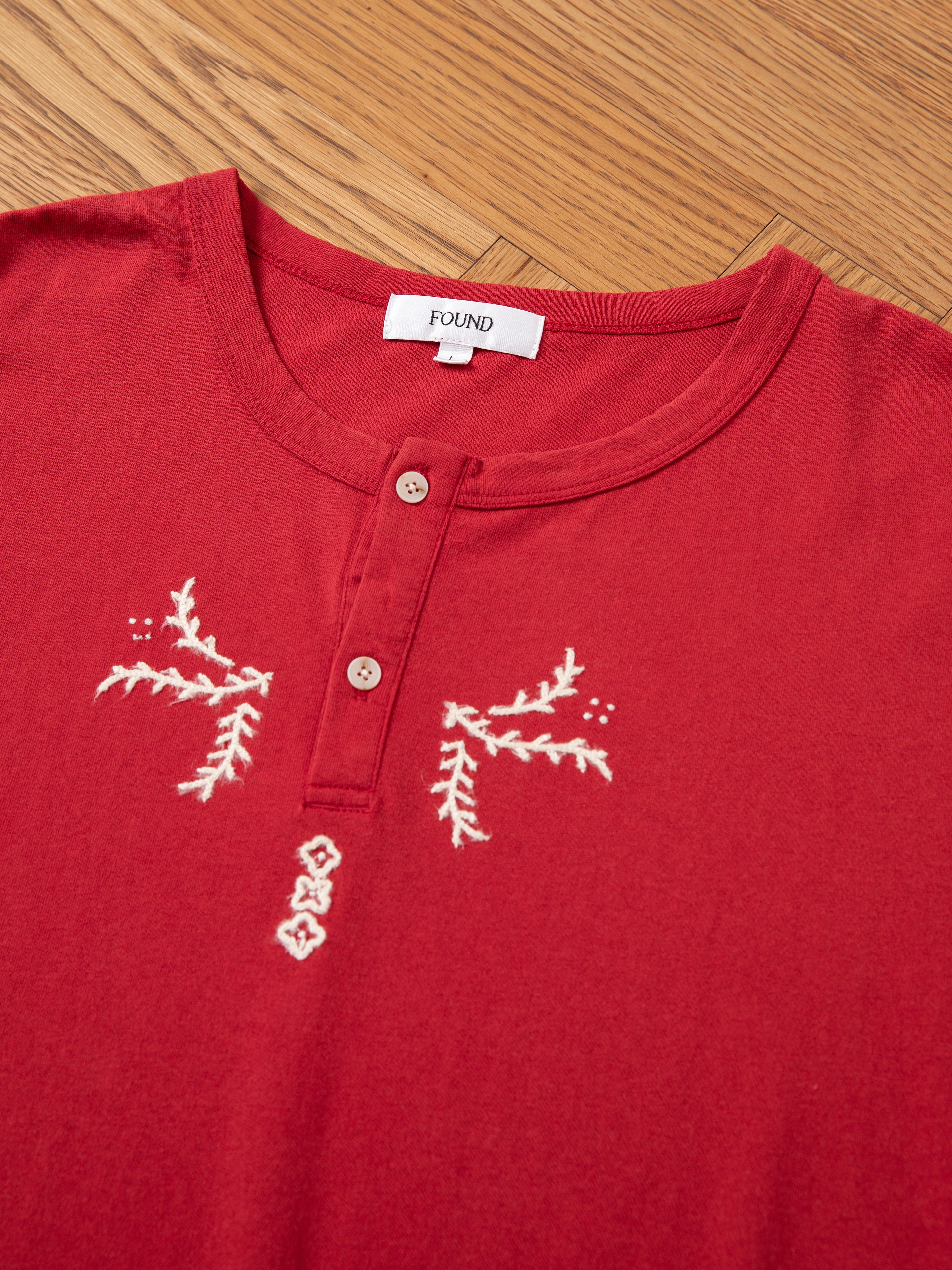 The Embroidered Henley by FOUND, a red shirt featuring white embroidery and a partially buttoned front, lies on a wooden surface. The label inside reads "FOUND.