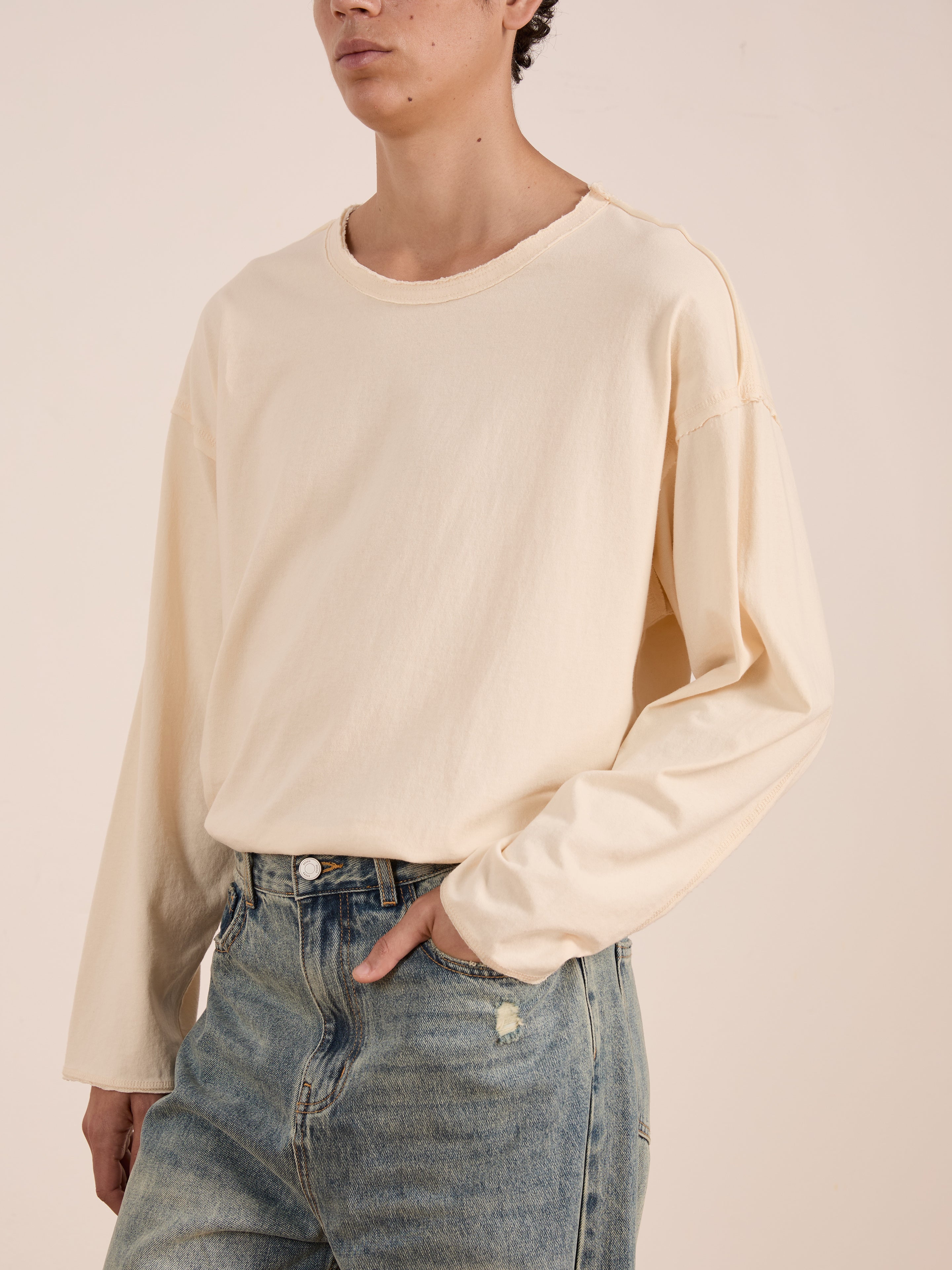 A person wears the FOUND Reversed LS Tee, a loose cream unisex long sleeve with a distressed neckline, paired with light blue jeans and stands with one hand in their pocket against a neutral background.