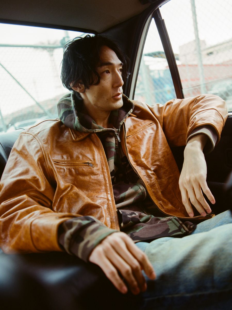 A man in a FOUND Marlboro Sun Faded Leather Racer Jacket and camouflage hoodie sits in the backseat of a car, gazing out the window.