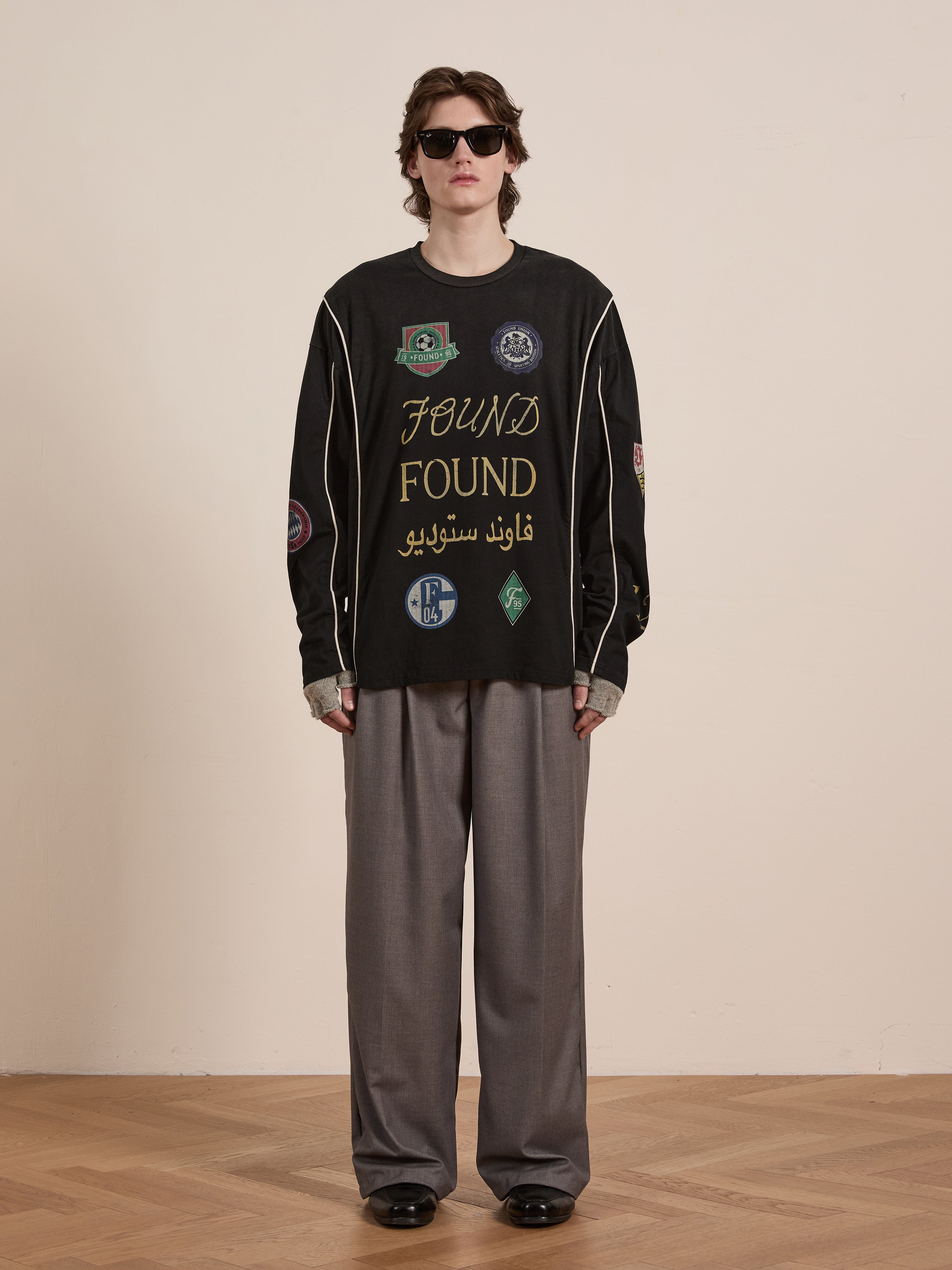 A person embodies unisex casual wear in a retro style, wearing sunglasses and the "FOUND" Multi Language LS Jersey in black, adorned with various patches. They pair it with gray wide-leg pants and black shoes while standing on a wooden floor against a plain wall.