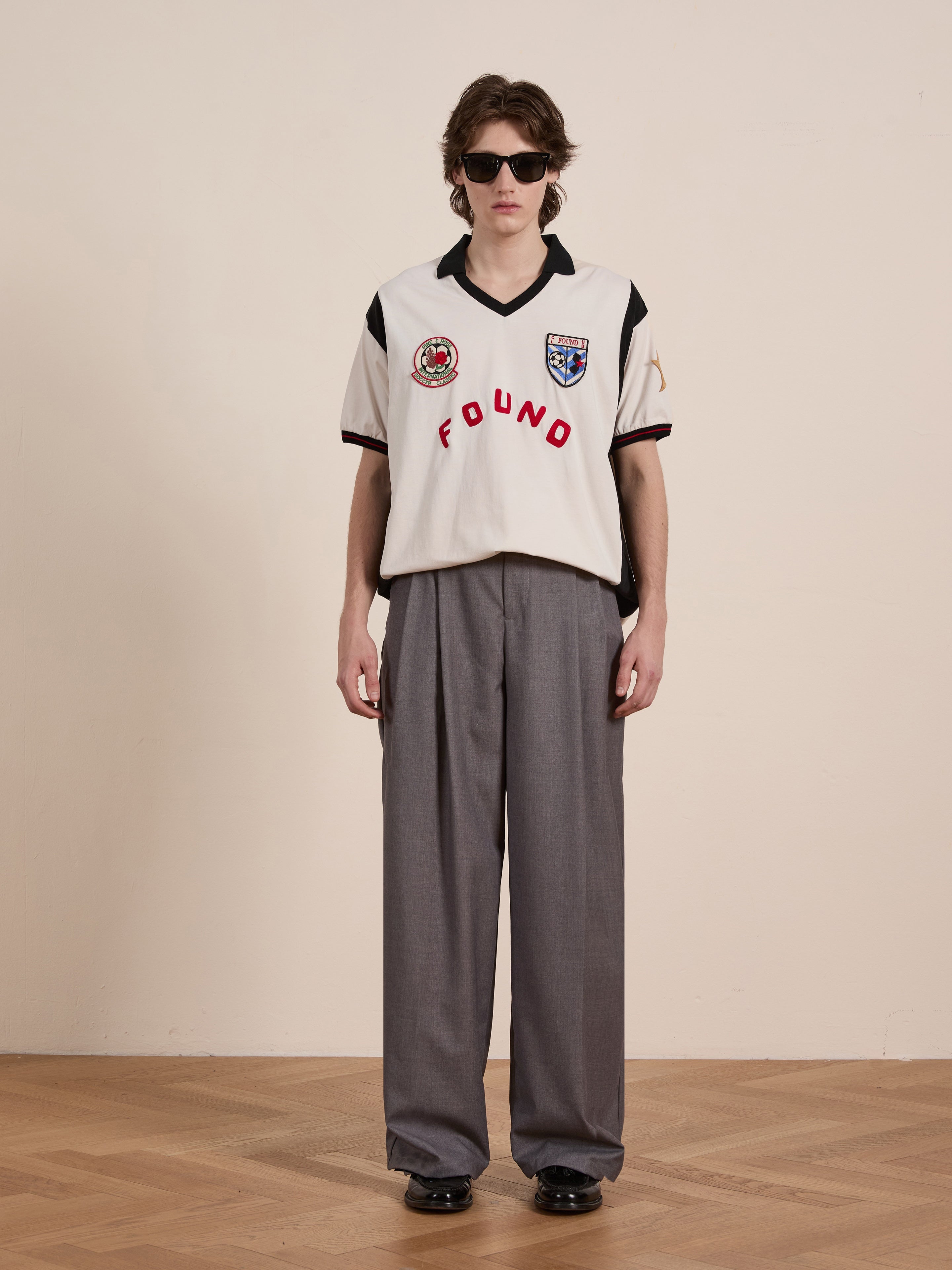 A person with a modern sportswear vibe stands on a wooden floor, wearing sleek sunglasses and FOUND's Union Soccer Jersey with patches, paired with loose gray trousers against a plain background.