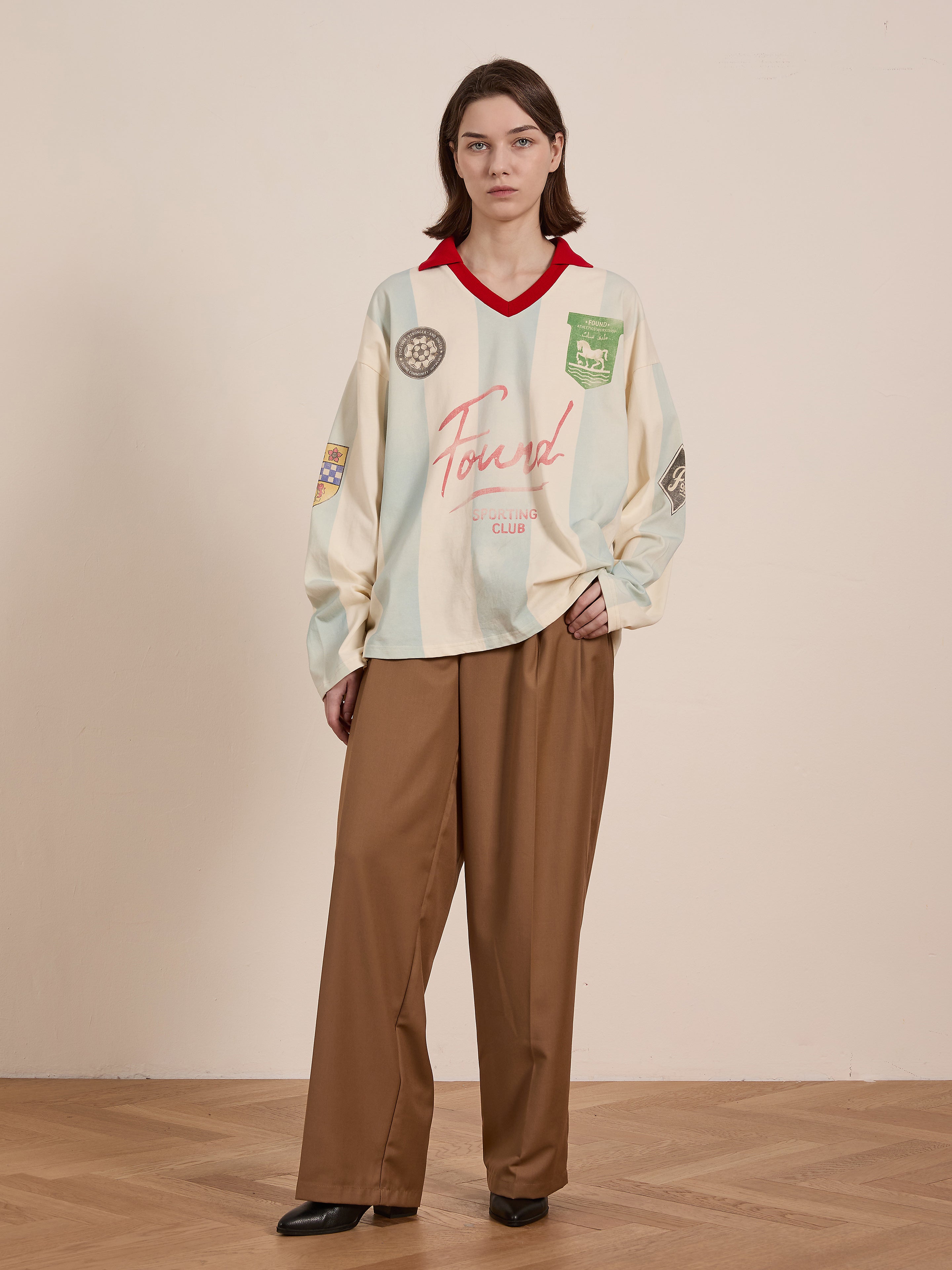 In a neutral-toned room, the individual wears the Stripe Sport LS Jersey by FOUND, which resembles a vintage football jersey. They pair it with brown wide-leg trousers and black shoes.