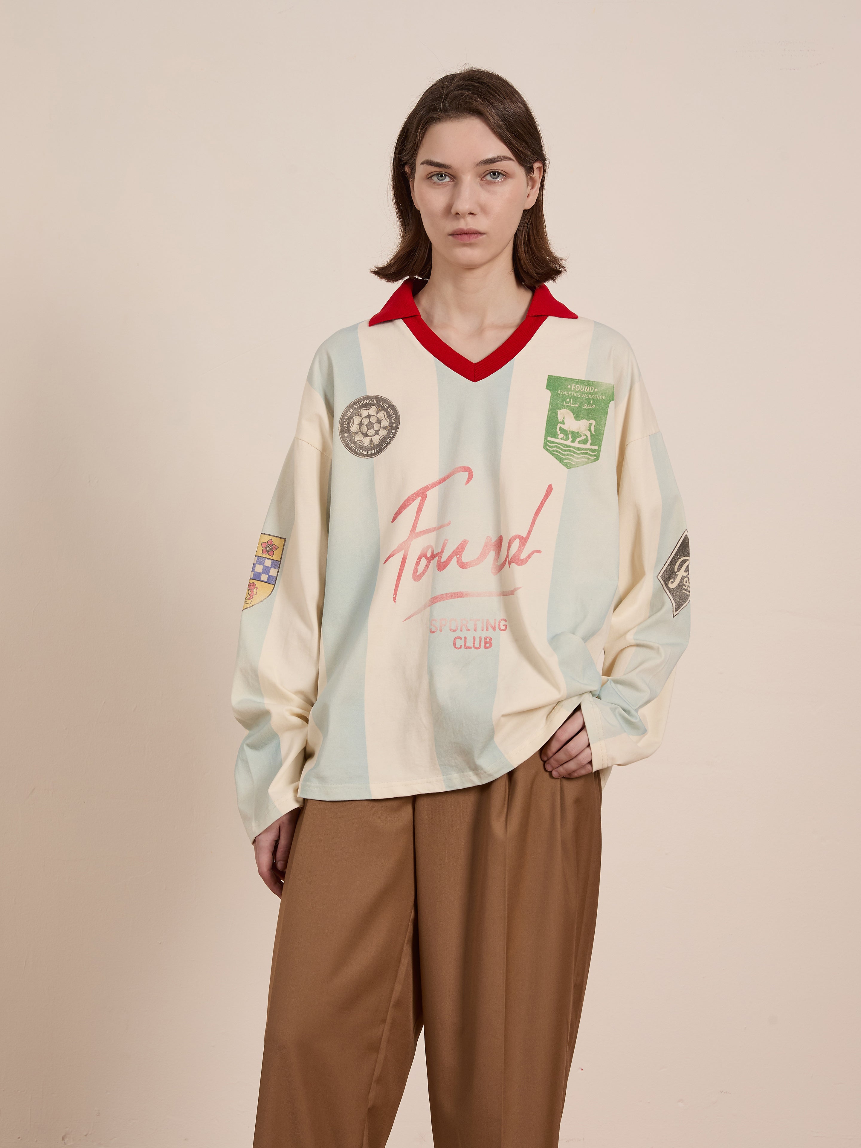 A person wearing the Striped Sport LS Jersey by FOUND, a cream and red vintage top with various patches, pairs it with brown pants while standing against a plain background.