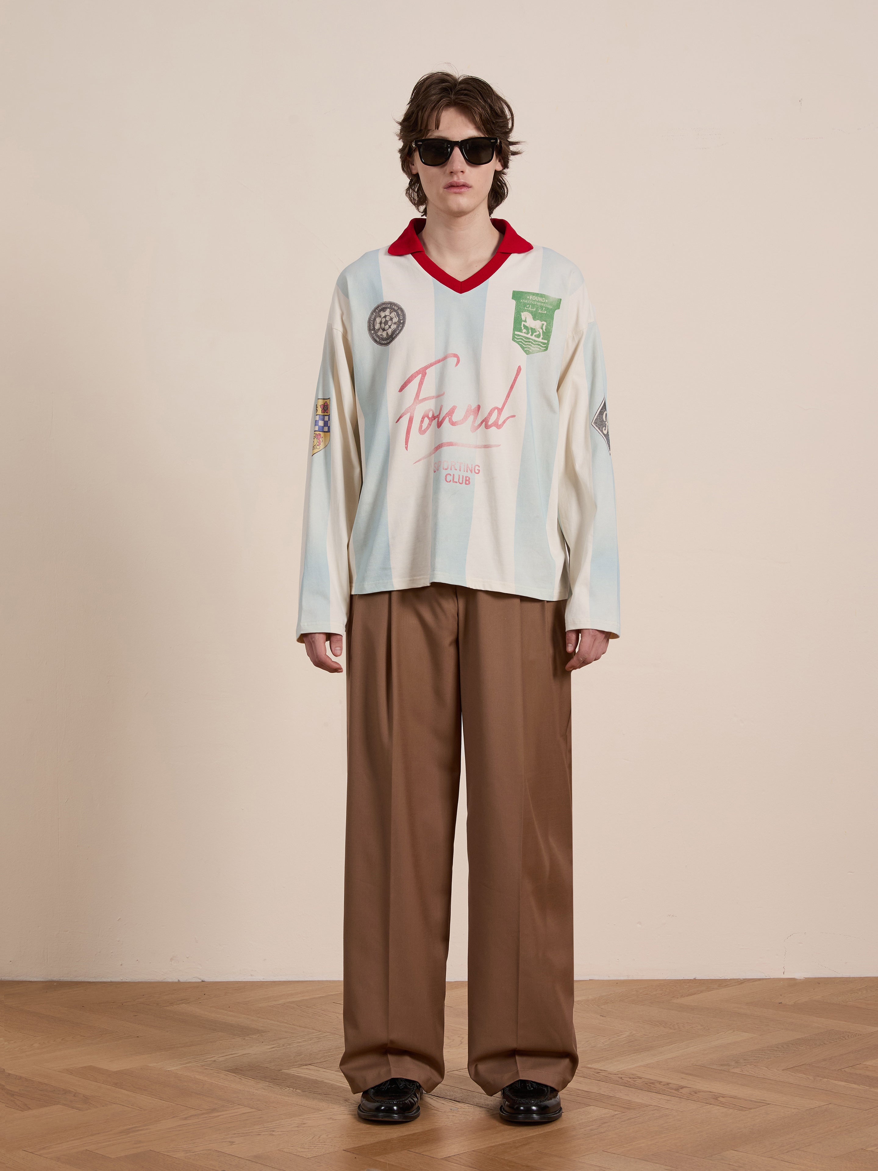 A person in sunglasses wears FOUND's Striped Sport LS Jersey, a light-colored vintage sportswear-inspired long-sleeve shirt with various patches, paired confidently with loose brown trousers on a wooden floor against a plain background.