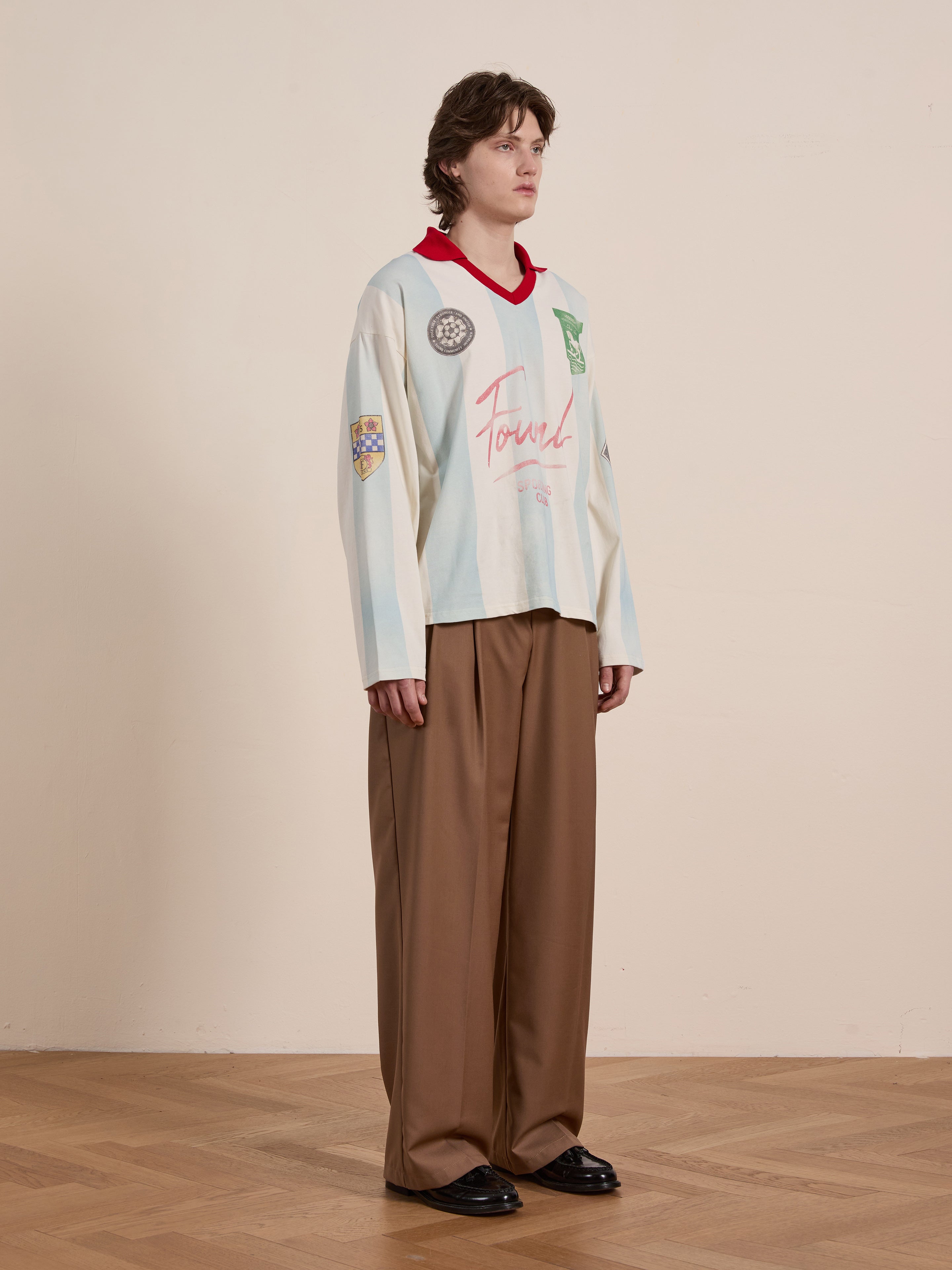 A person stands against a light background wearing FOUND's heritage-inspired Stripe Sport LS Jersey: a loose-fitting white long sleeve shirt with patches, paired with brown wide-leg pants and black shoes.