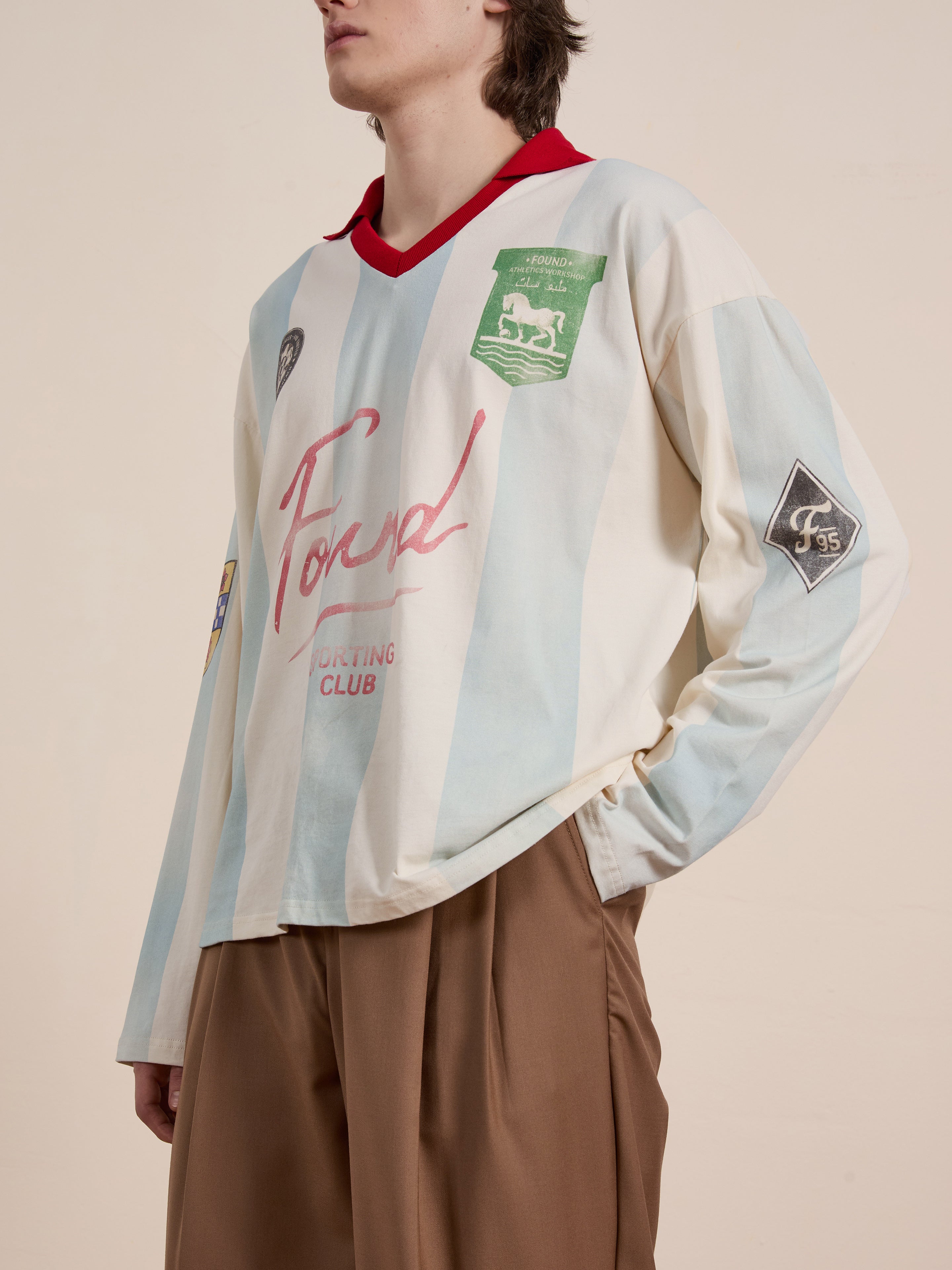 Wearing the FOUND Stripe Sport LS Jersey, featuring a red collar and sponsor logos, paired with brown pants creates a standout vintage sportswear look against a neutral background. This light blue and white unisex relaxed fit ensemble exemplifies classic style.