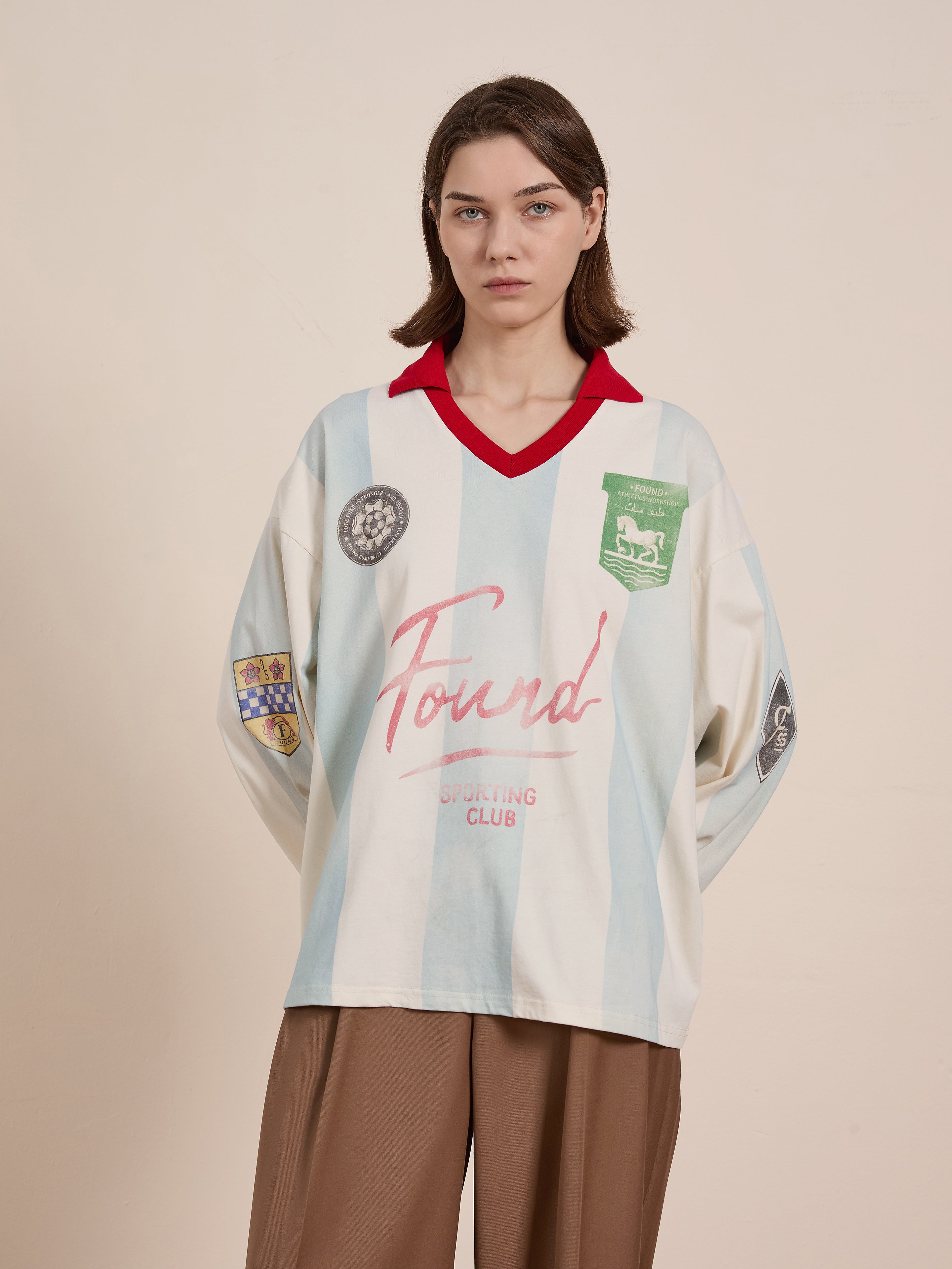 A person wears the Stripe Sport LS Jersey by FOUND, featuring light blue and white stripes with a red collar, various patches, and "Found Sporting Club" text. Styled with brown pants, it offers a unisex relaxed fit inspired by heritage sportswear.