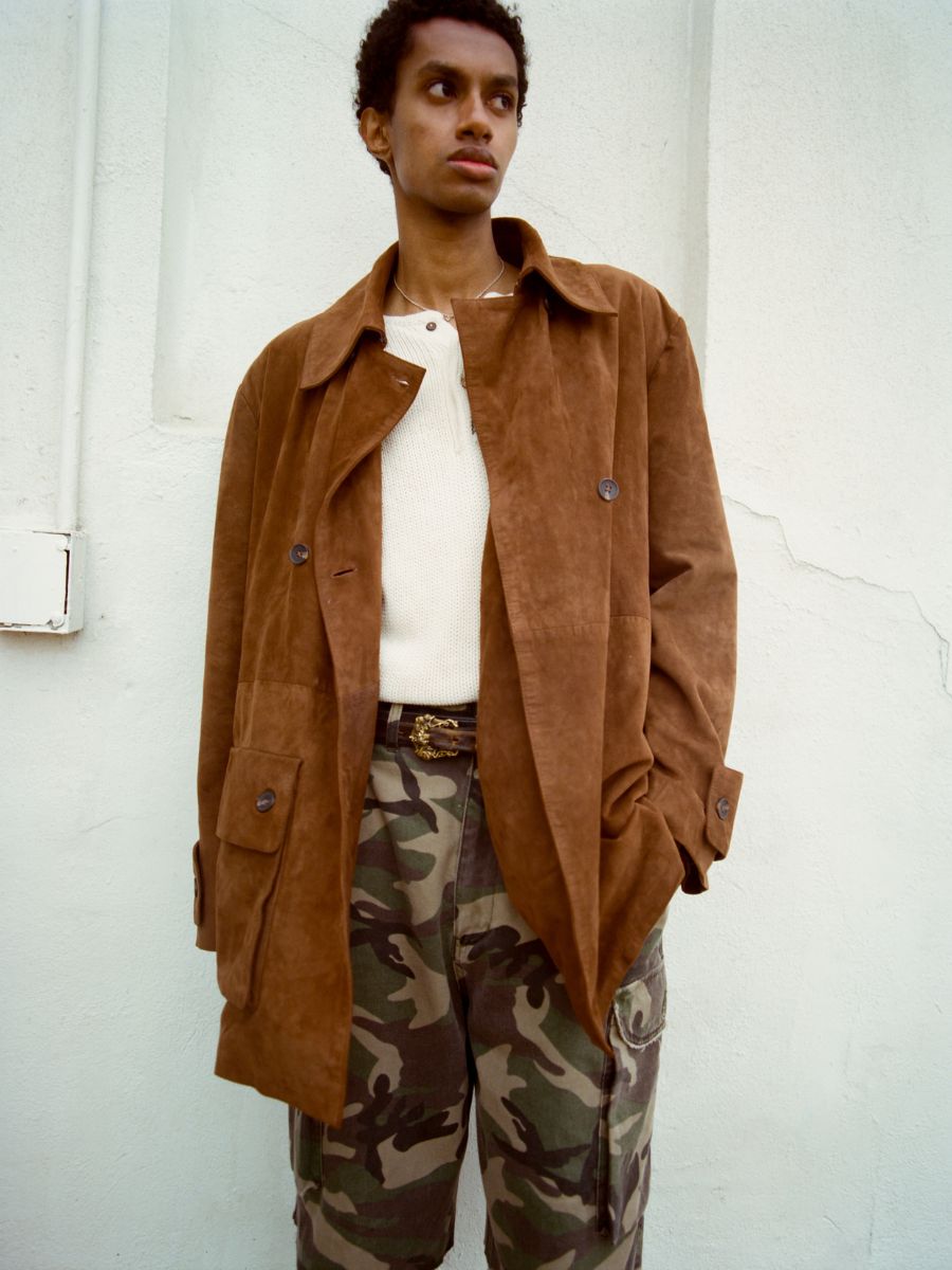 A person wears the FOUND Double Breasted Suede Leather Jacket over a white shirt and camouflage pants, standing against a light-colored wall.