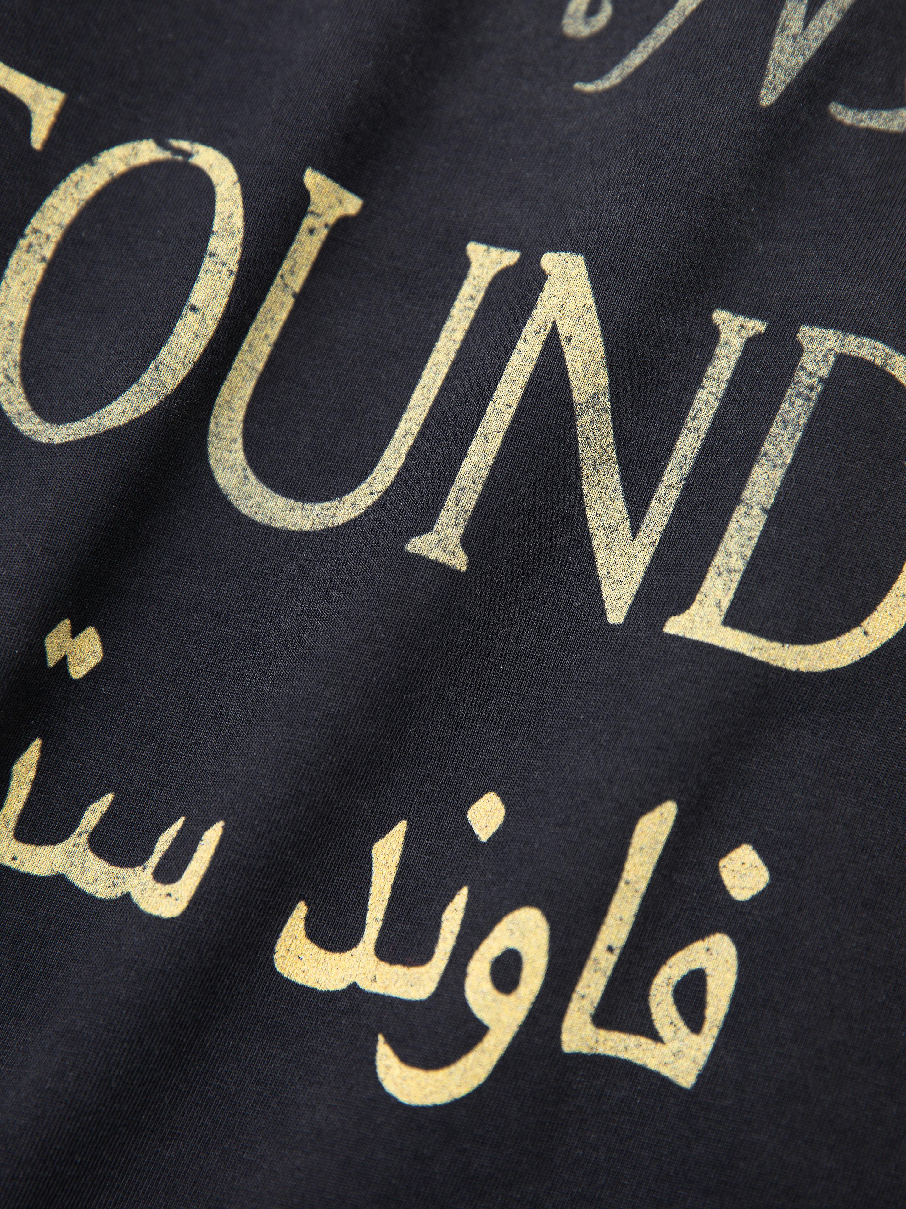 Close-up of a black fabric with a retro design, showcasing "FOUND" in worn gold lettering and Arabic script below—ideal for lovers of unisex casual wear with a vintage touch. This is the Multi Language LS Jersey by FOUND.