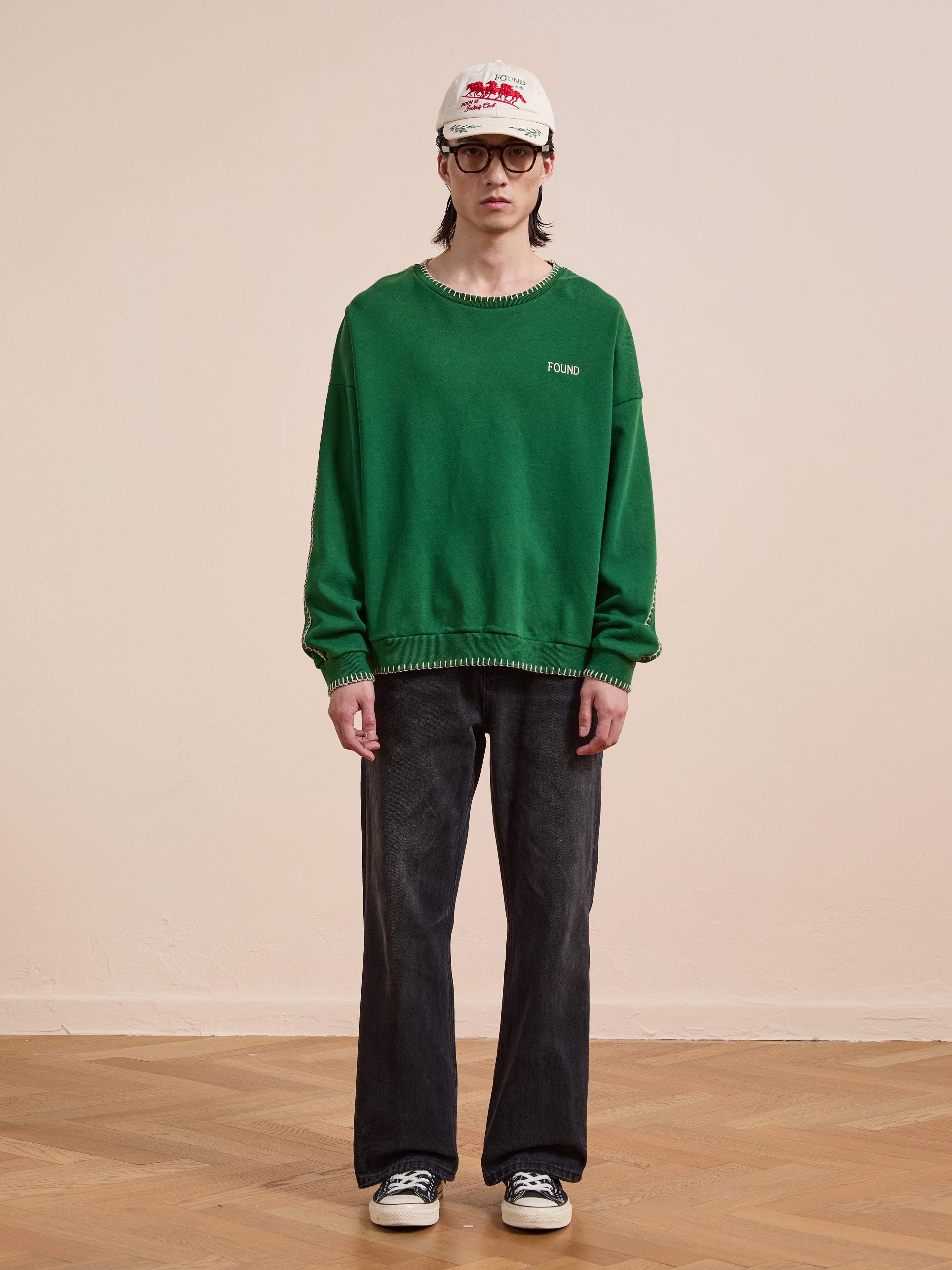 Someone wearing the FOUND Stitched Border Distressed Crewneck, dark jeans, black and white sneakers, glasses, and a white cap stands indoors on a wooden floor against a plain beige wall.