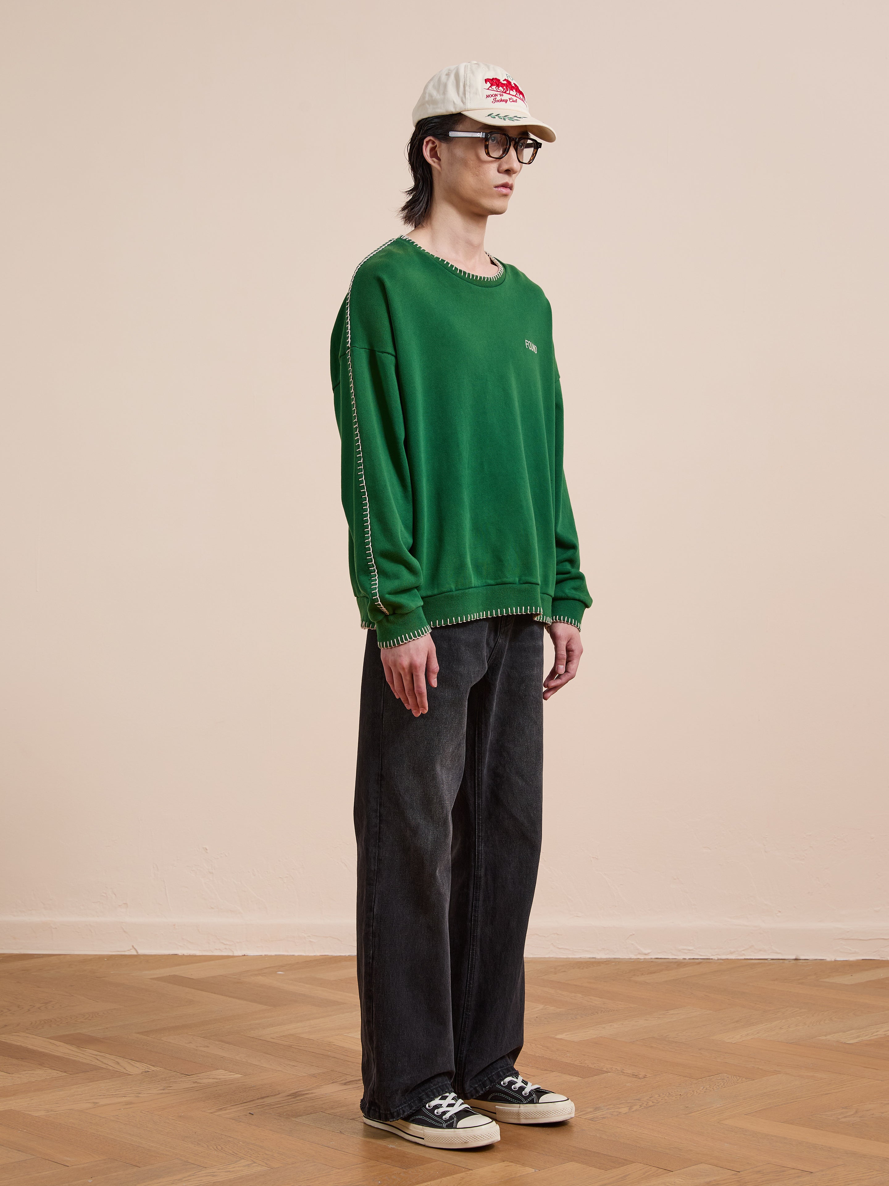 A person stands indoors against a beige wall, wearing FOUND's Stitched Border Distressed Crewneck in green with a white cap, glasses, wide-leg dark jeans, and black sneakers on a wooden floor.