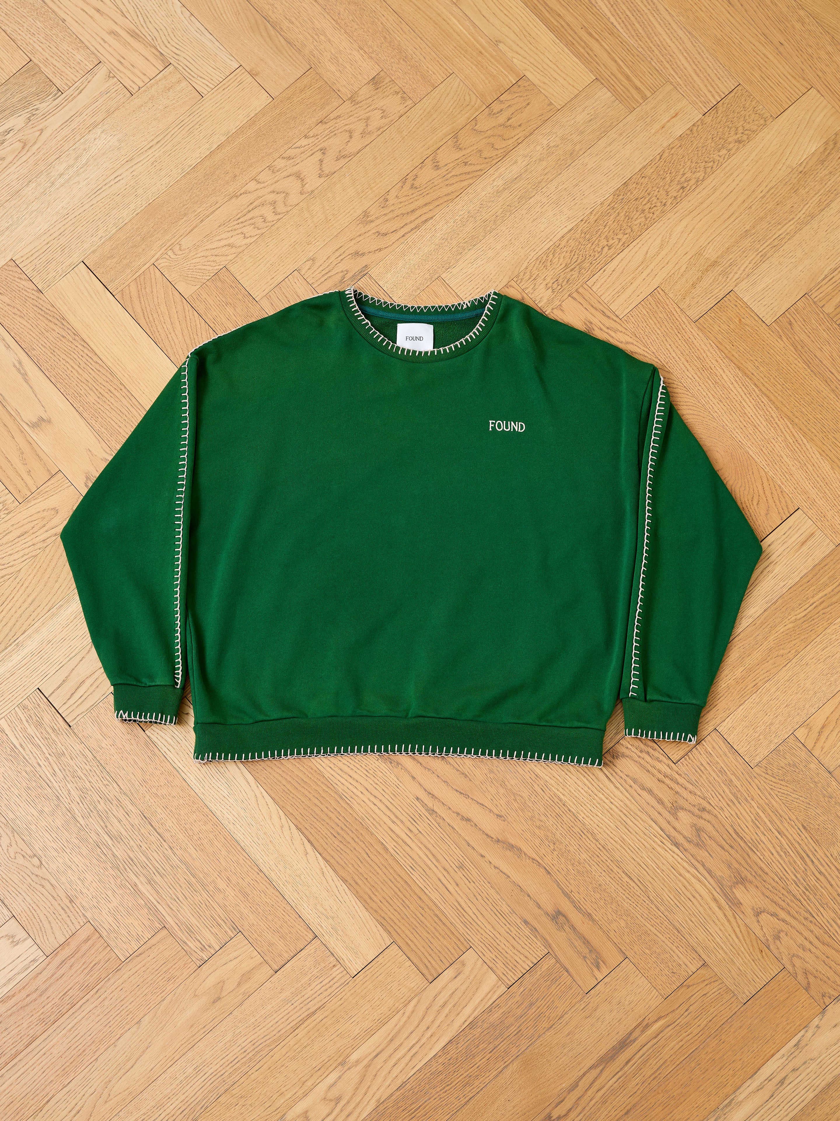 The FOUND Stitched Border Distressed Crewneck, a green vintage-style top with white stitching and "FOUND" embroidered on the chest, is displayed flat on a wooden herringbone floor.