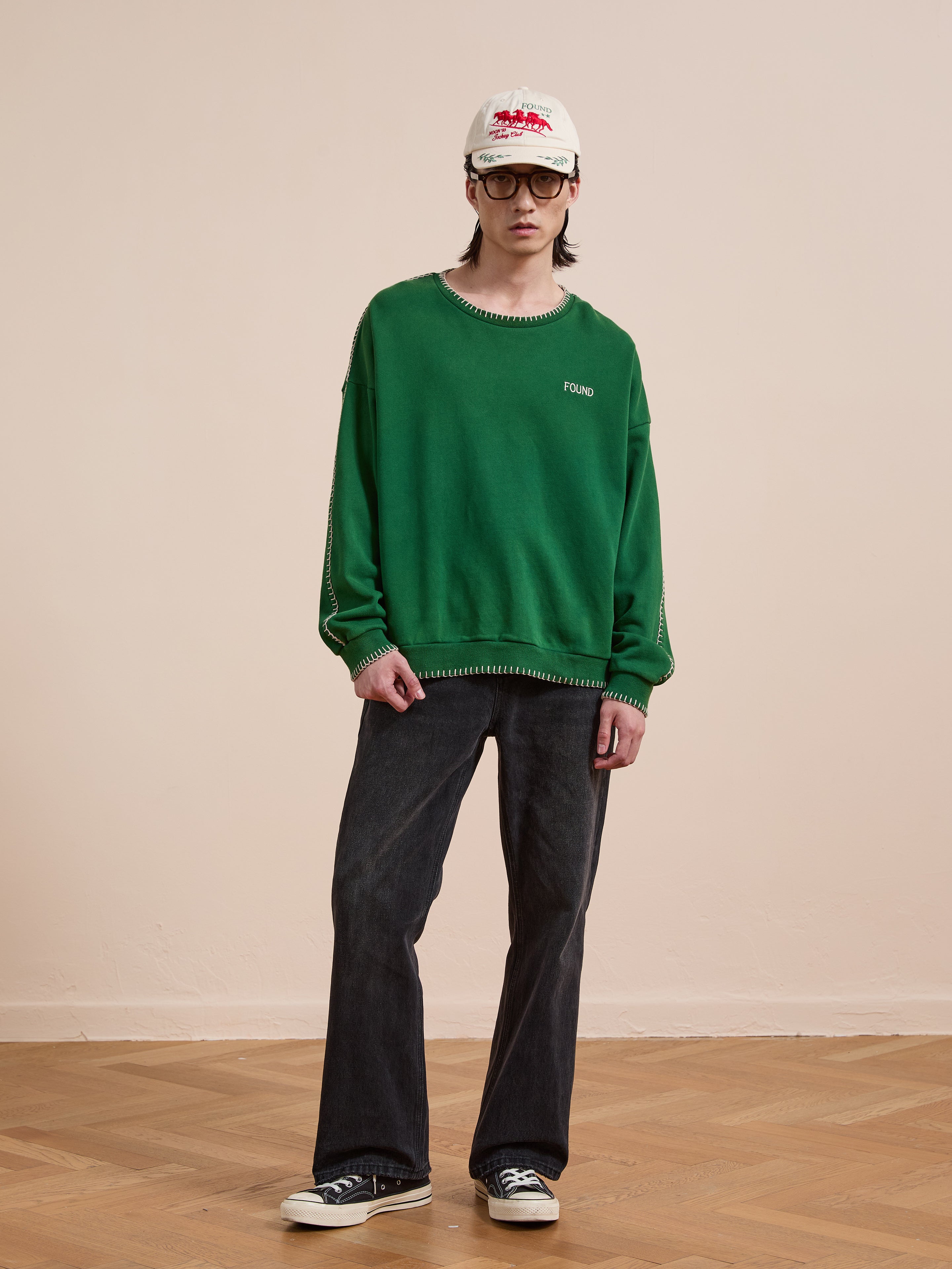 A person stands indoors on a wooden floor against a plain beige wall, wearing the FOUND Stitched Border Distressed Crewneck, black jeans, black sneakers, a white cap, and glasses.